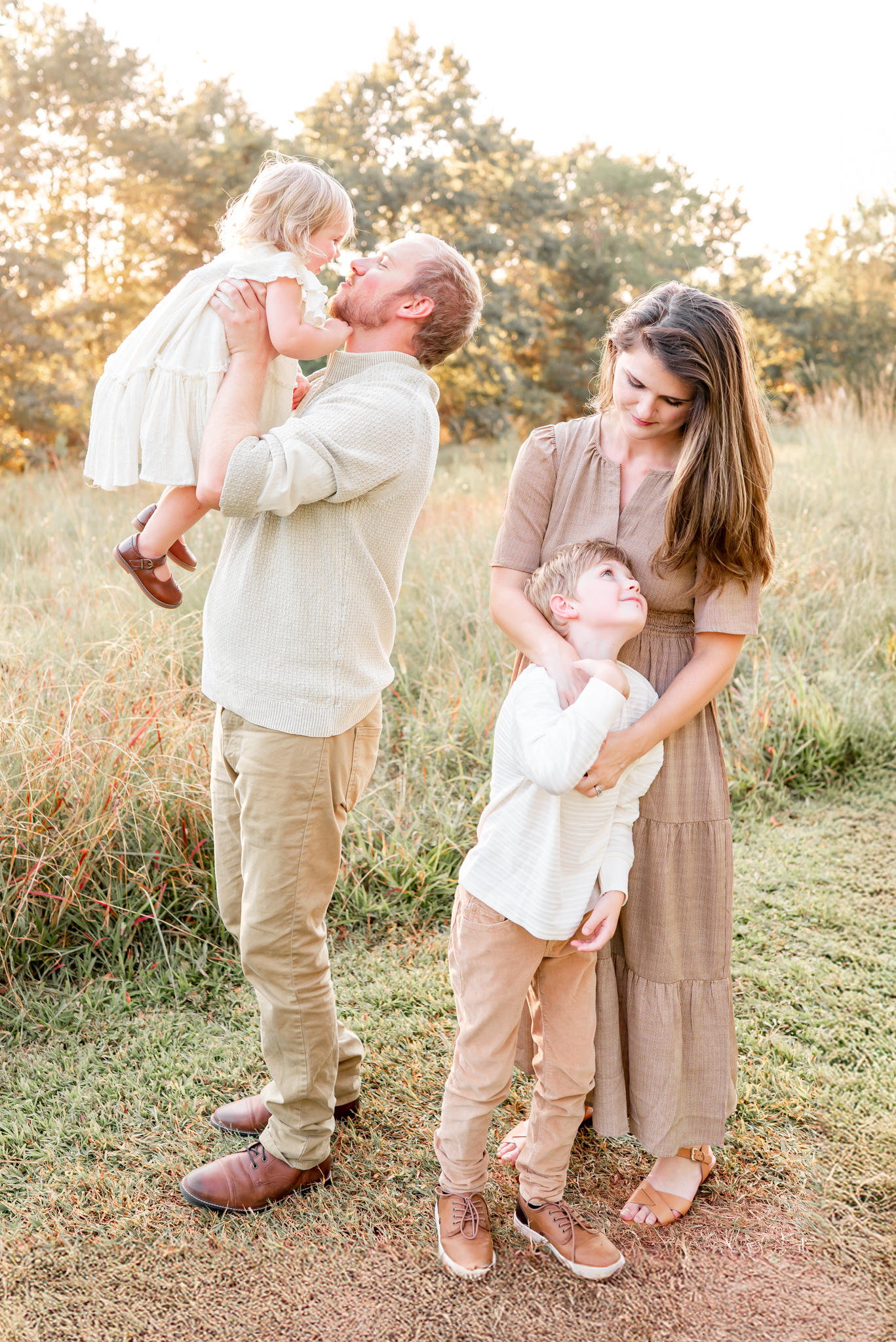 A family of 4, dressed in whites, khakis, and browns in a grassy field. The dad is holding the toddler daughter in the air. The mom and school age son are hugging off to the side.