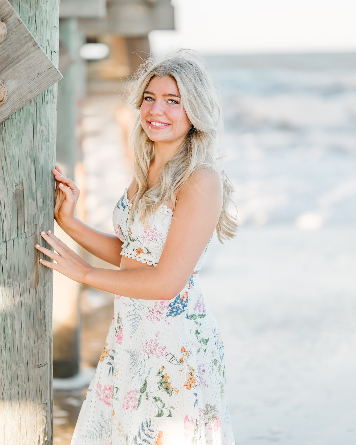 A high school senior in a floral skirt and top leans against a pier at the beach. The water can be seen behind her.