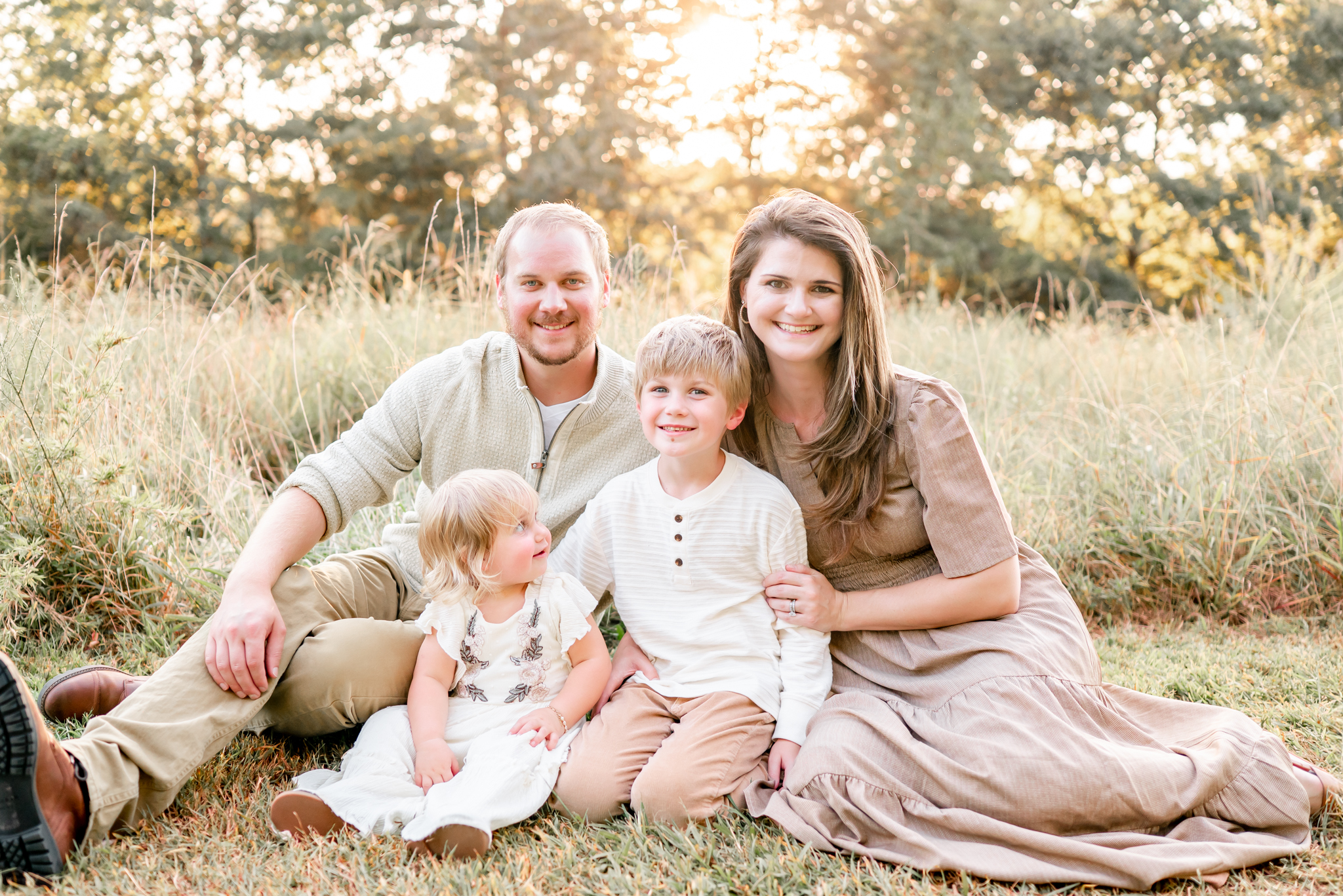 A family of 4 sits in the grass. The sun is shining in the trees behind them. They are dressed in neutral outfits.