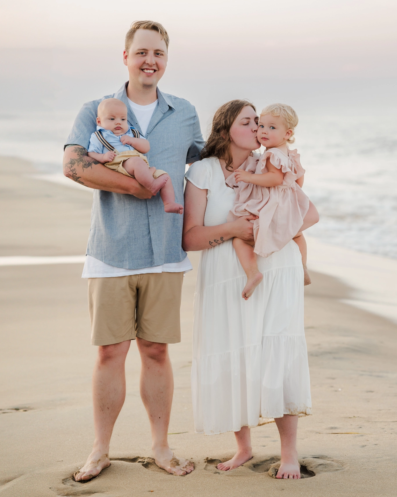 A family of 4 stands together in front of the waves at the beach. Sunset is turning the sky behind them shades of pink and purple. They are dressed in blues, whites, and khakis.