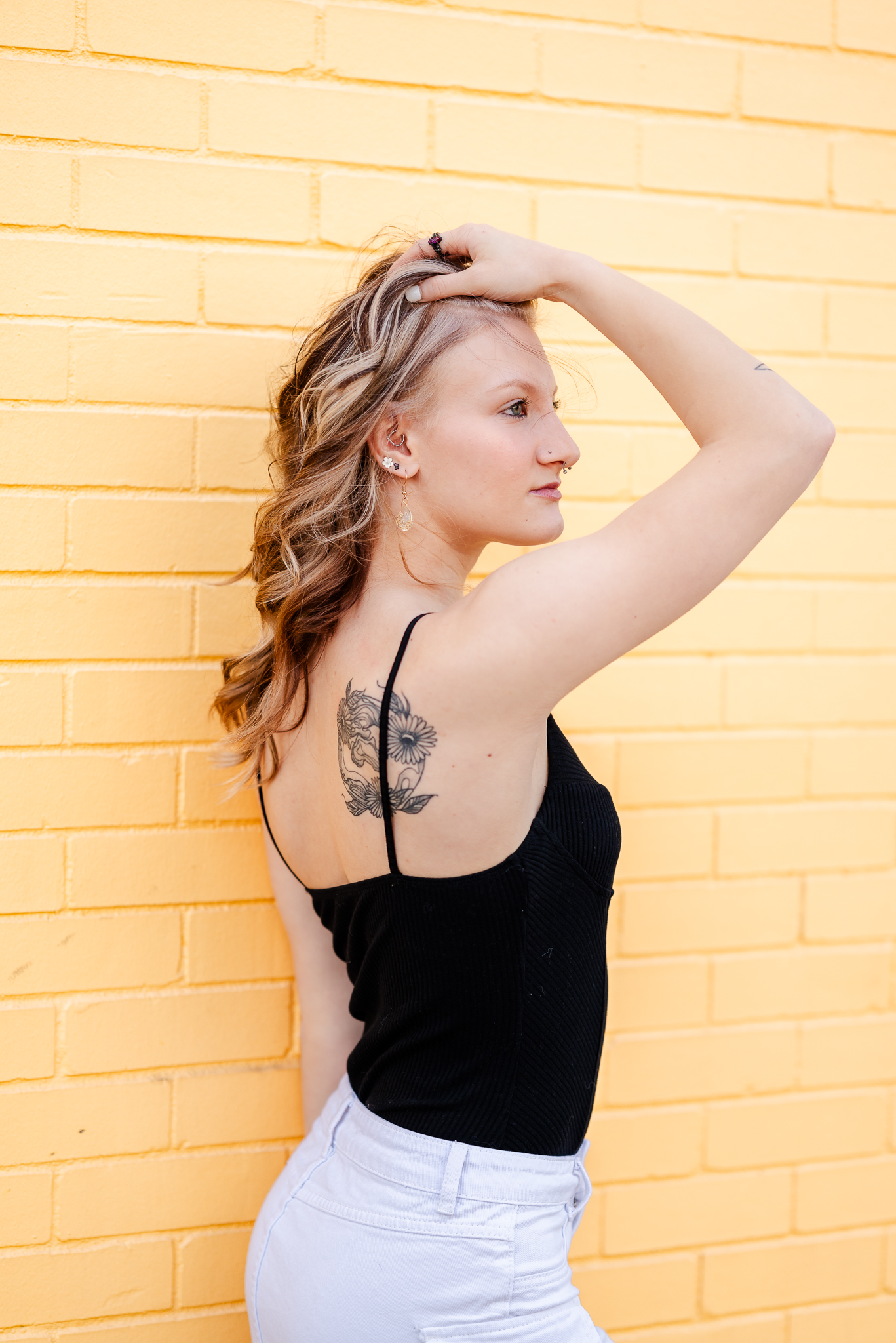 A high school senior shows off the tattoo on her shoulder. She is holding her hair up and looking off into the distance. There is a yellow, painted brick wall behind her.