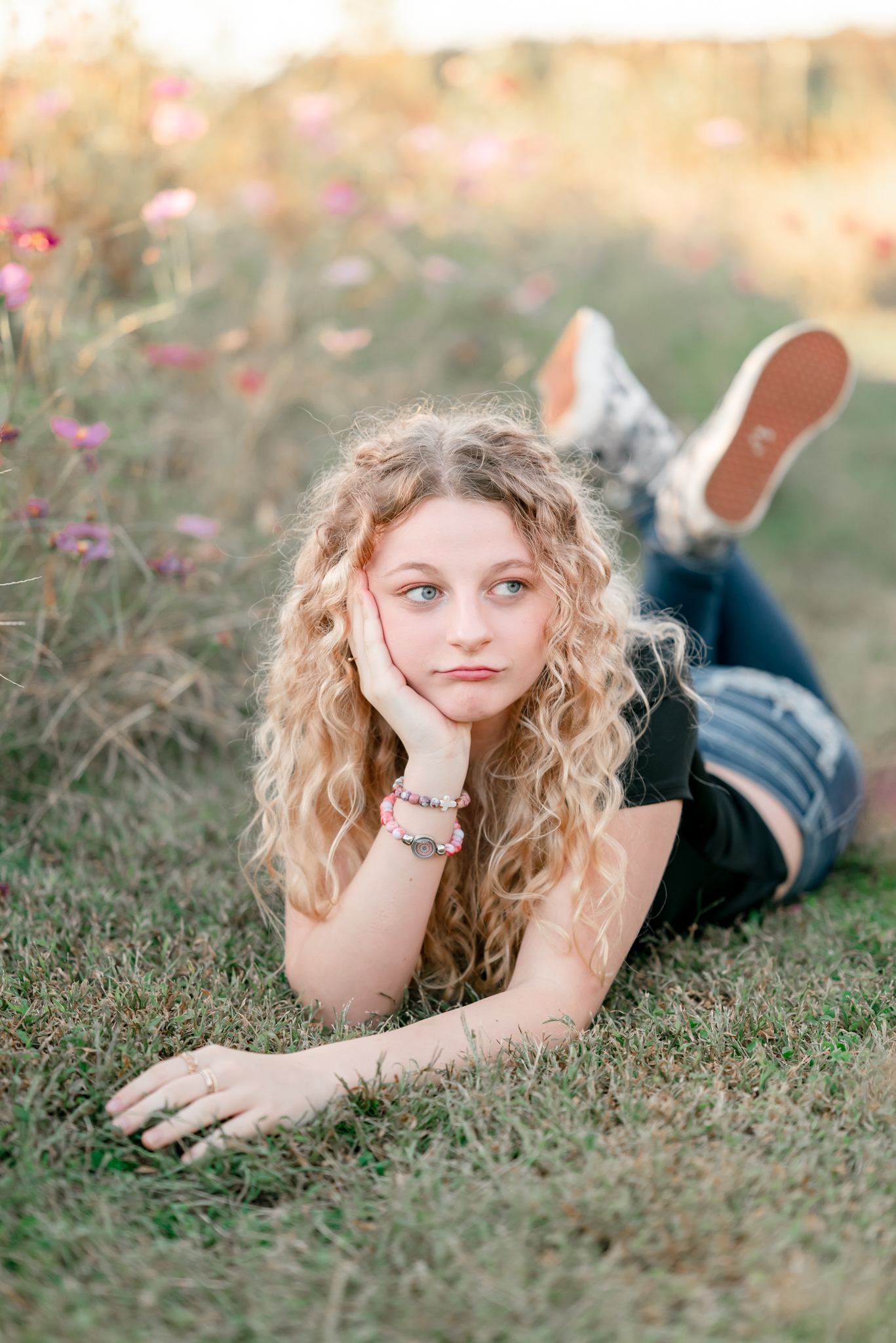 A high school senior, wearing a black t-shirt and jeans sits on her belly in a field of wildflowers. She rests her chin in her hands and looks off to the side with a quirky expression on her face.