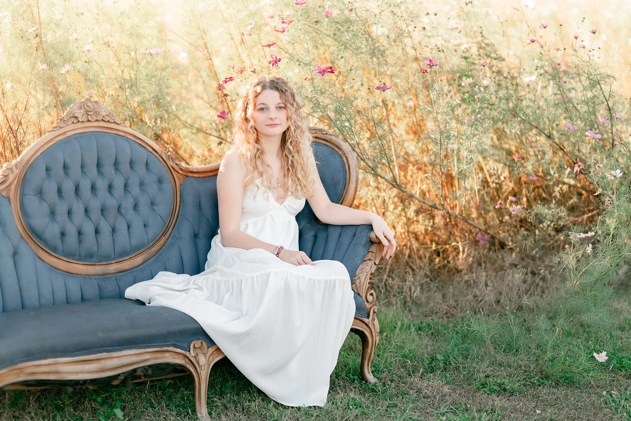 A senior in a white dress, sits on an antique blue couch among the flowers. Image captured at Backyard Wildflower Patch in Chesapeake, VA.