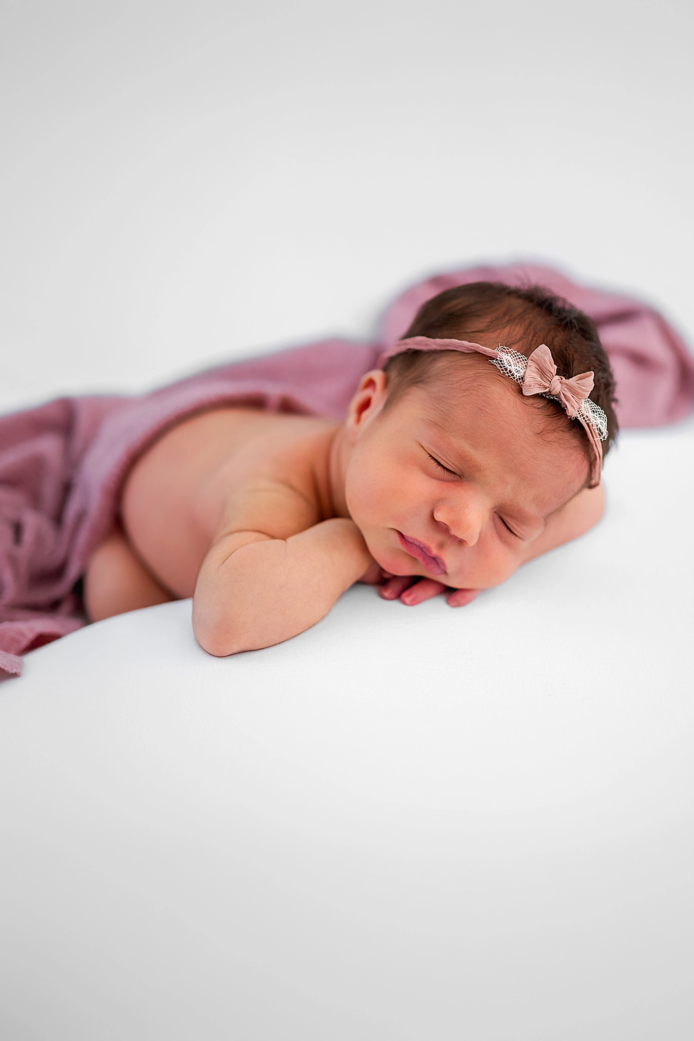 A newborn baby girl sleeps on a white blanket. She has a mauve blanket over her and a matching headband in her hair. Image captured by Chesapeake Newborn photographer, Justine Renee Photography.