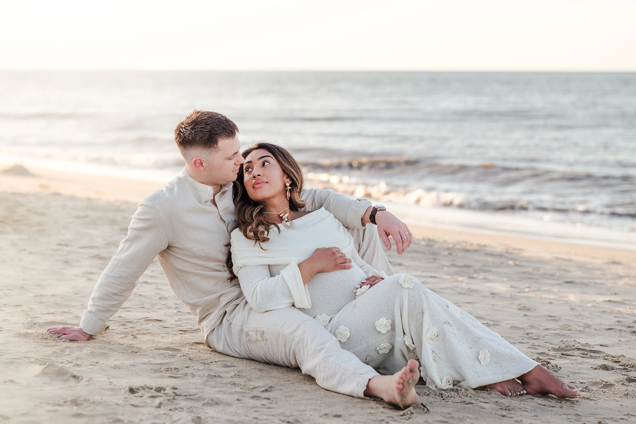 An expectant mother in a beige sweater dress, leans against her husband, who is also in neutral colors. Captured by Chesapeake maternity photographer, Justine Renee Photography.