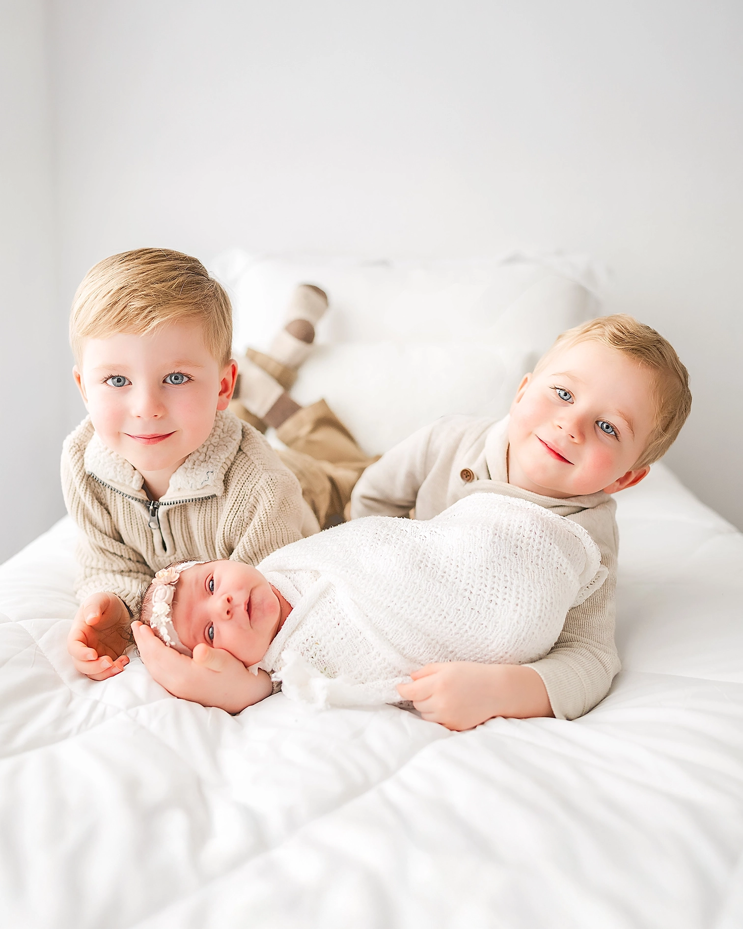 A pair of toddler boys in beige lay on their stomachs on a bed. Their newborn sister, swaddled in white, lays between them.