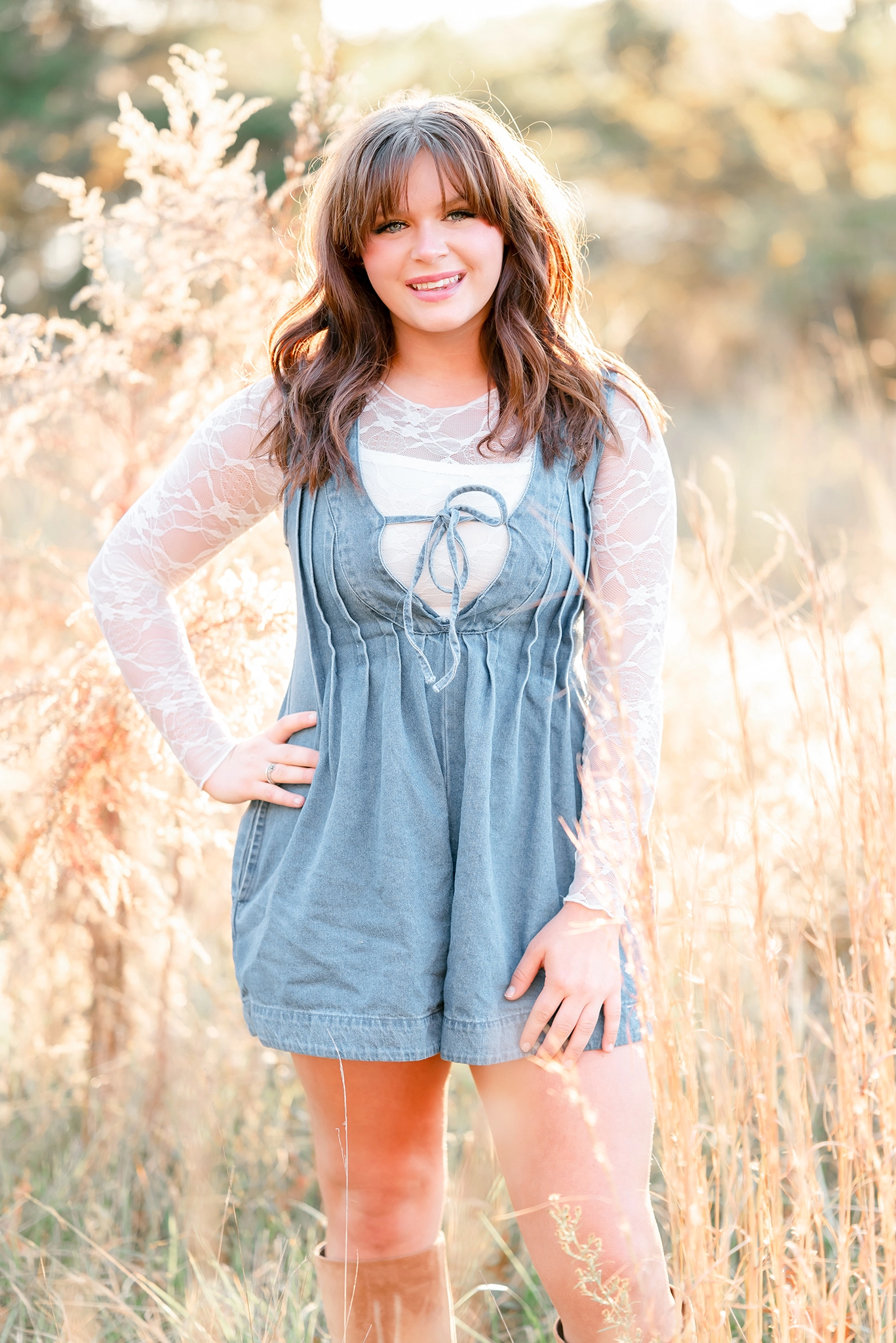A high school senior in a lace top and denim romper smiles big while standing in a field of tall grass. She is having her senior portraits taken by Chesapeake photographer, Justine Renee Photography