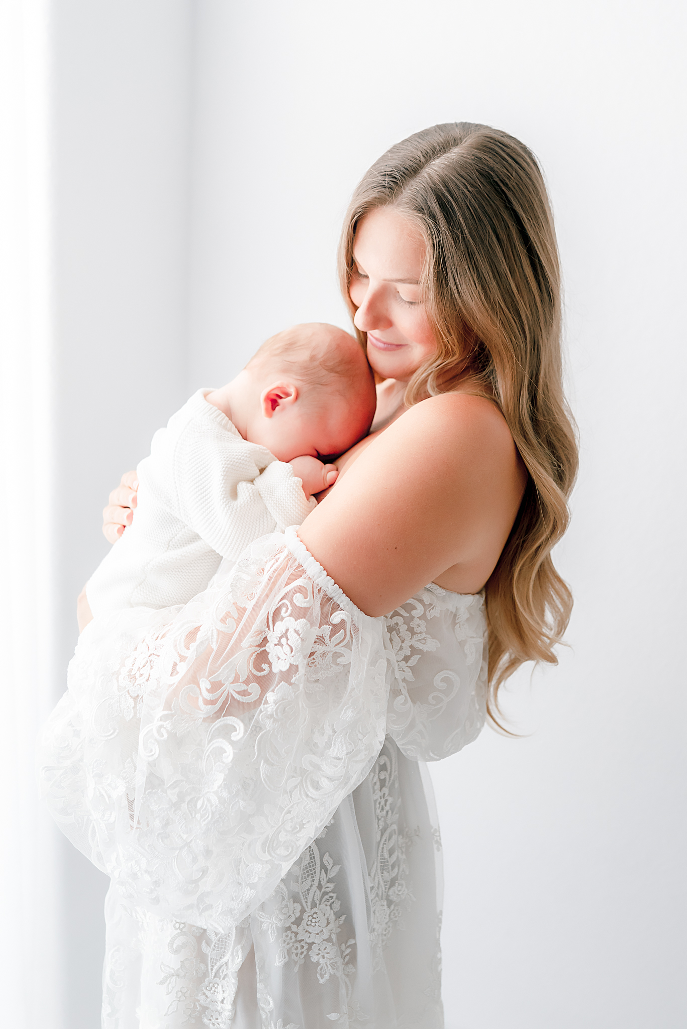 A young mother, holding her newborn baby, captured by Chesapeake photographer, Justine Renee Photography. The mother, is dressed in a white lace dress available in Justine's client closet. Her baby is snuggle up to her chest.