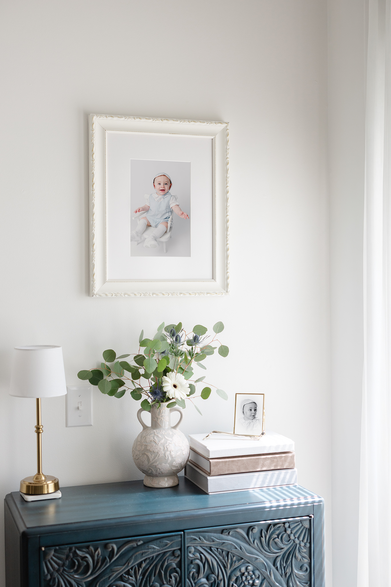 An heiroom portrait hangs on the wall of a home's entry way. The boy is wearing a white and blue outfit with matching bonnet. One the table below sits a black and white profile of the same young boy.