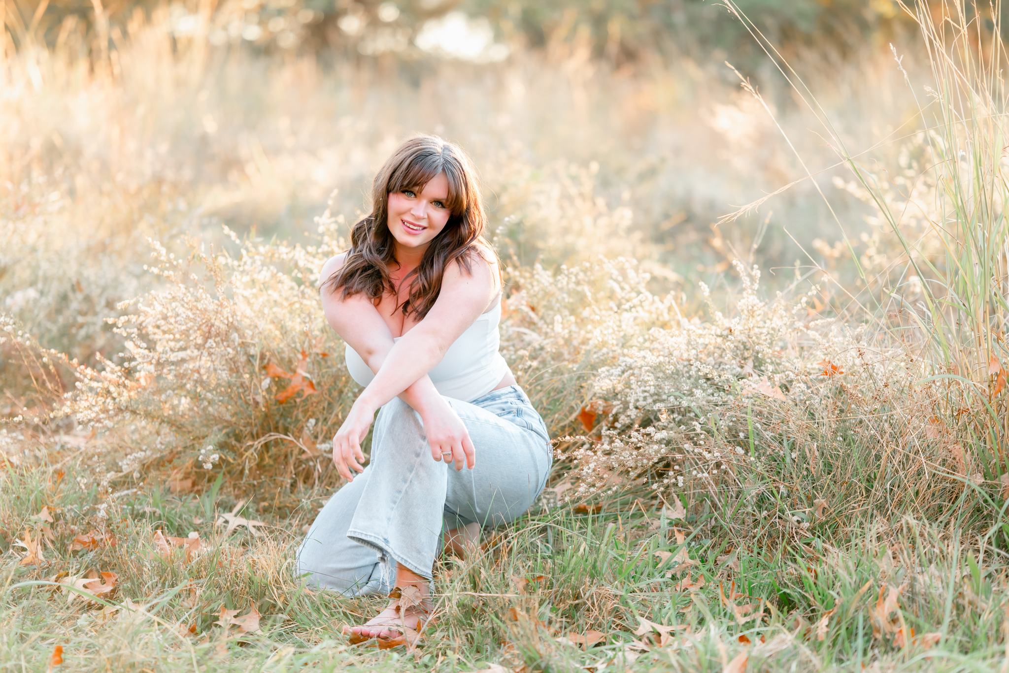 A high school senior wearing jeans and a white t-shirt kneels in some tall grass. The grass at Bells Mill Park in Chesapeake always looks golden in the fall. A high school senior wearing jeans and a white t-shirt kneels in some tall grass. The grass at Bells Mill Park in Chesapeake always looks golden in the fall.