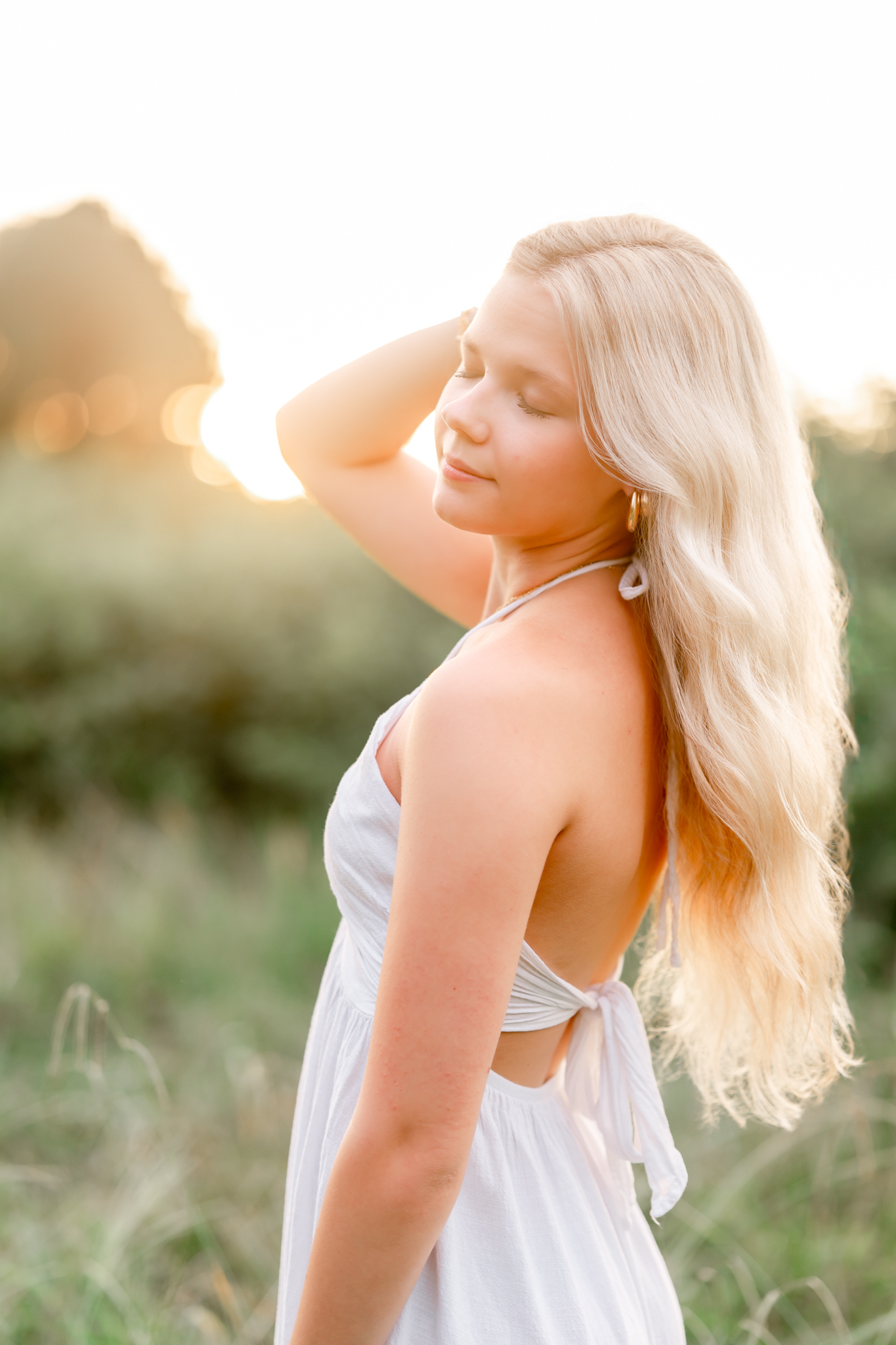 A high school senior, in a white dress, tosses her hair over her should as she closes her eyes. The sun in shining through the trees behind her and the tall grass is green and wild.