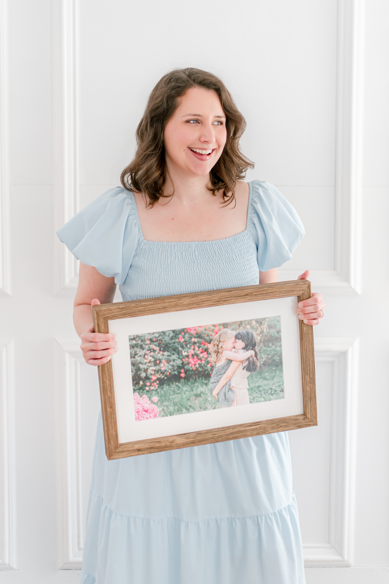 Chesapeake photographer, Justine Renee Photography is pictured. She is wearing a a blue dress and standing in front of a white wall. She is holding a framed picture of her girls, loving on each other.