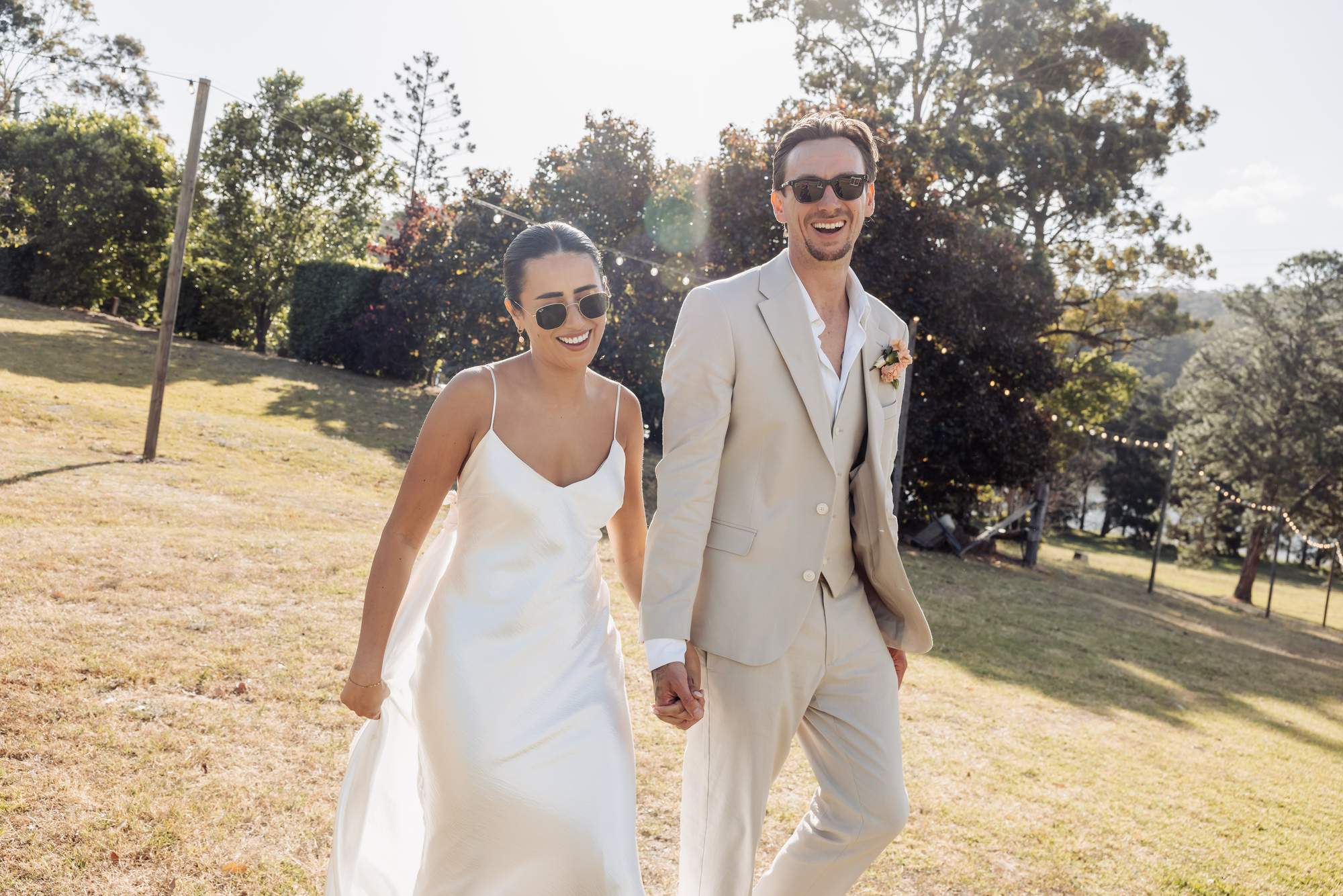 Newlyweds in sunglasses walk hand in hand across a sunlit lawn