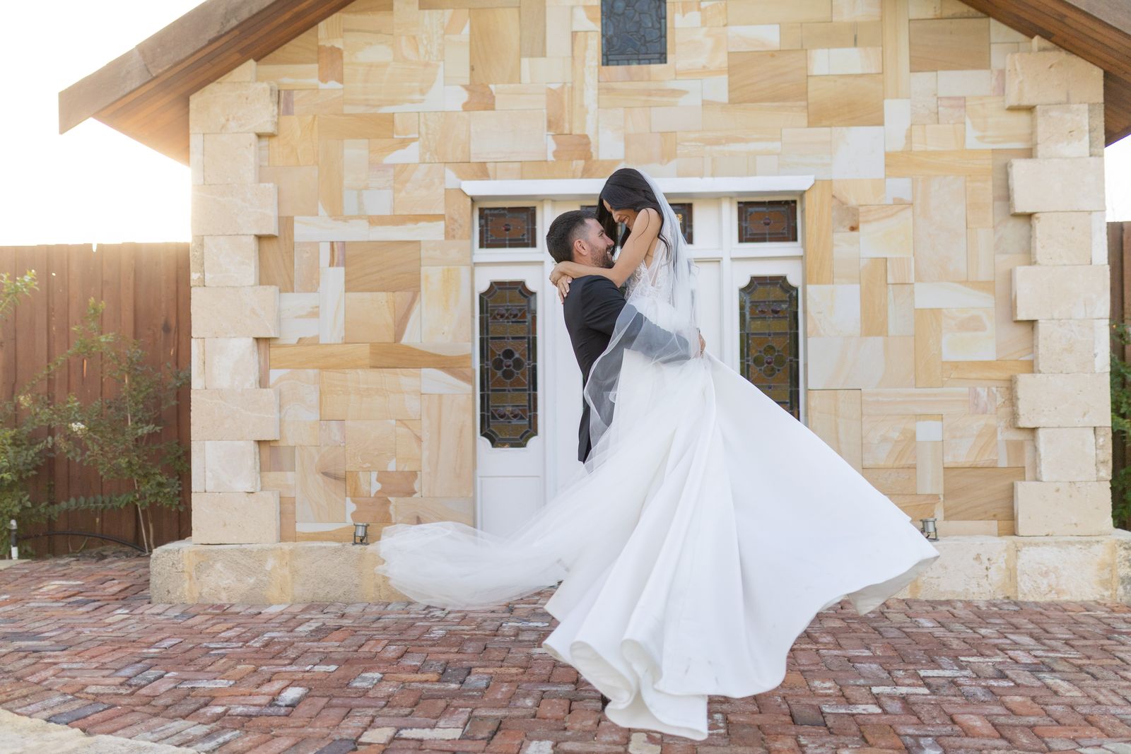 Groom lifts his bride outside a sandstone chapel, her dress flying