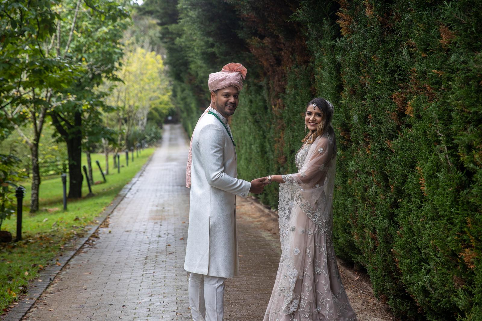Groom in a sherwani holds hands with his bride on a garden path