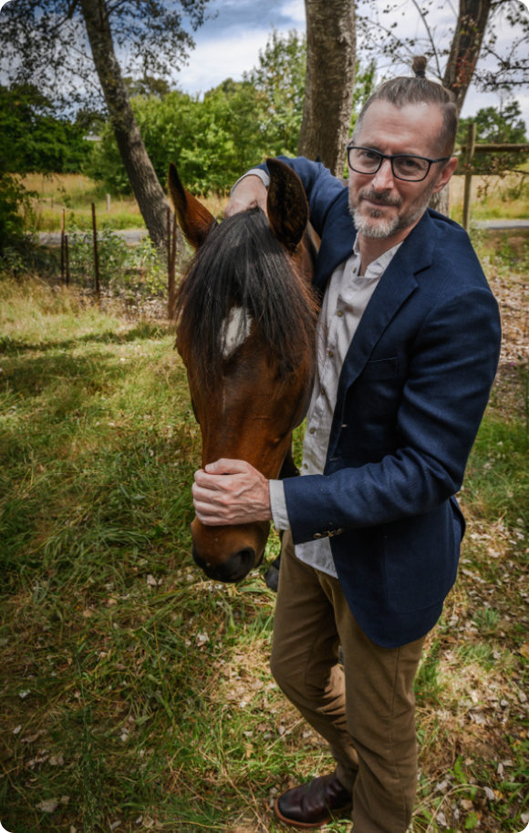 Photo Of Dr. Ed Standing With A Horse, His Right Hand Holding The Horse's Neck Behind The Ears, His Left Hand Resting On The Horse's Nose Just Above The Nostrils