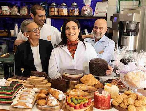 A medium-shot photograph of the Settepani family—Leah Abraham, Nino Settepani, and their children Bilena and Seyoum—smiling behind a wooden counter filled with an abundant display of Italian pastries and artisanal desserts. Bilena stands at the center in a white chef’s coat and patterned scarf, resting her hand on a large, dark panettone. Leah sits to the left in a black blazer and glasses, Seyoum stands behind her in a white t-shirt, and Nino stands to the right in a light blue button-down shirt. The foreground is crowded with colorful treats, including rainbow cookies, cannoli, fruit tarts, croissants, and various cakes, while shelves of glass jars and coffee equipment are visible in the background of their Harlem bakery.