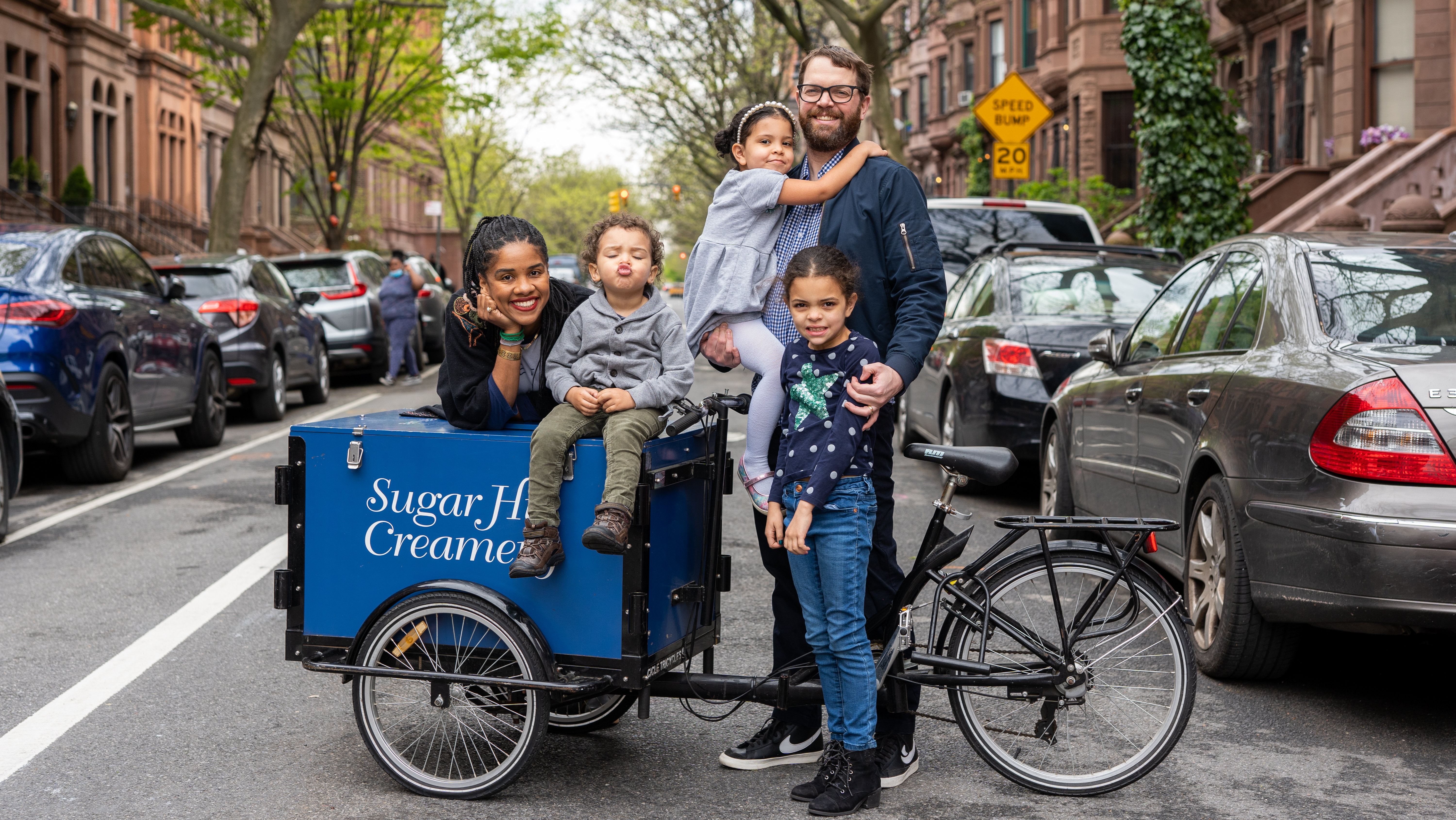A bright outdoor photograph by Ajene Farrar features Petrushka Bazin Larsen, her husband Nick Larsen, and their three children on a brownstone-lined Harlem street. The family is posed around a blue Sugar Hill Creamery branded delivery tricycle; Petrushka leans against the cargo box while their young son sits on top, and Nick stands nearby holding one daughter while the other stands in front. This image captures the family's deep community roots and their entrepreneurial spirit within the neighborhood they have called home for 20 years.