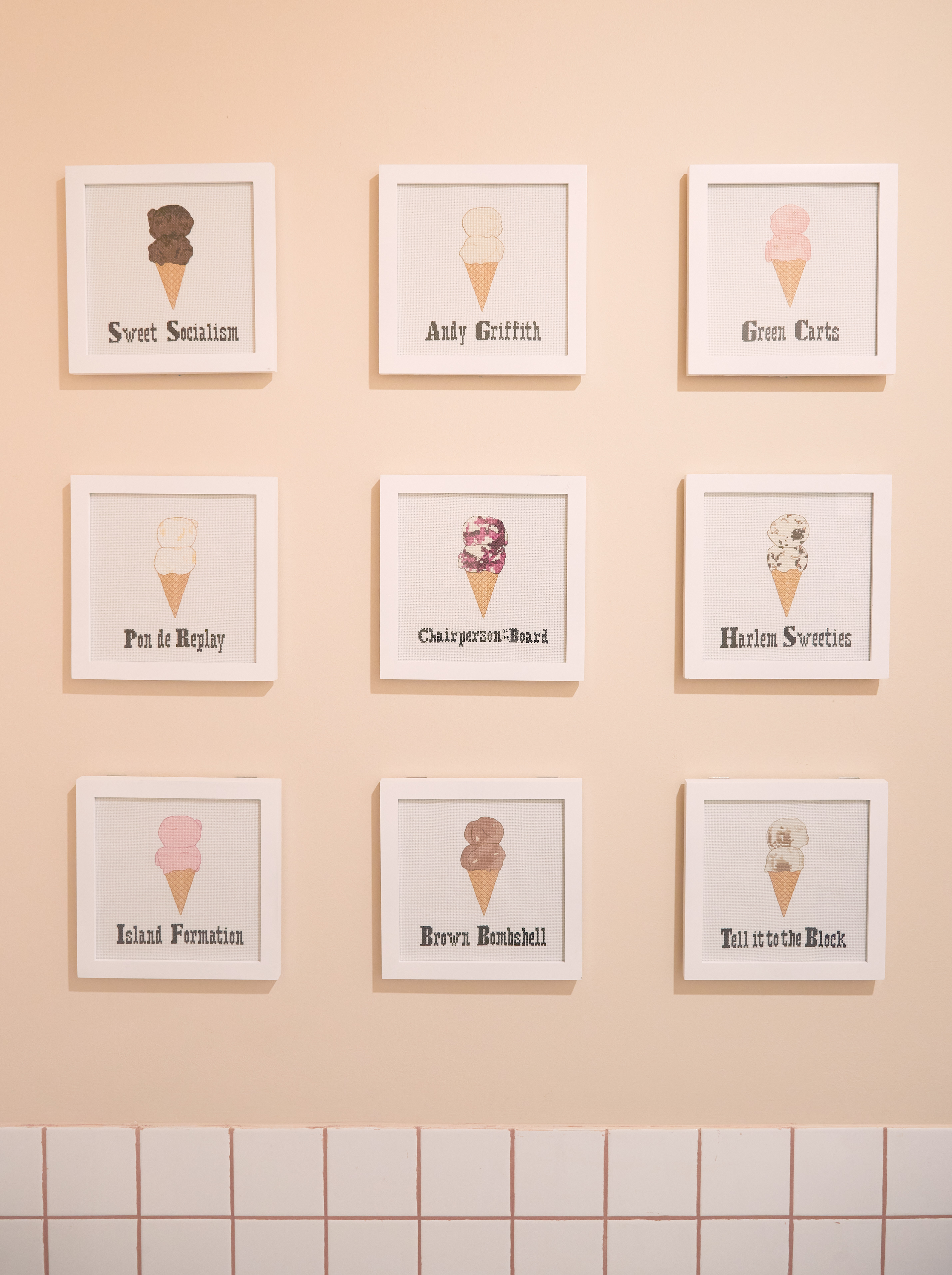 A photograph by Perry Hall features nine square white frames arranged in a three-by-three grid on a light peach-colored wall above white tiling. Each frame contains a minimalist illustration of an ice cream cone with two scoops, labeled with a unique, culturally-inspired flavor name from Sugar Hill Creamery. The featured flavors include:  Sweet Socialism (dark chocolate sorbet)  Andy Griffith (vanilla)  Green Carts  Pon de Replay  Chairperson of the Board (blueberry cheesecake)  Harlem Sweeties  Island Formation  Brown Bombshell  Tell it to the Block