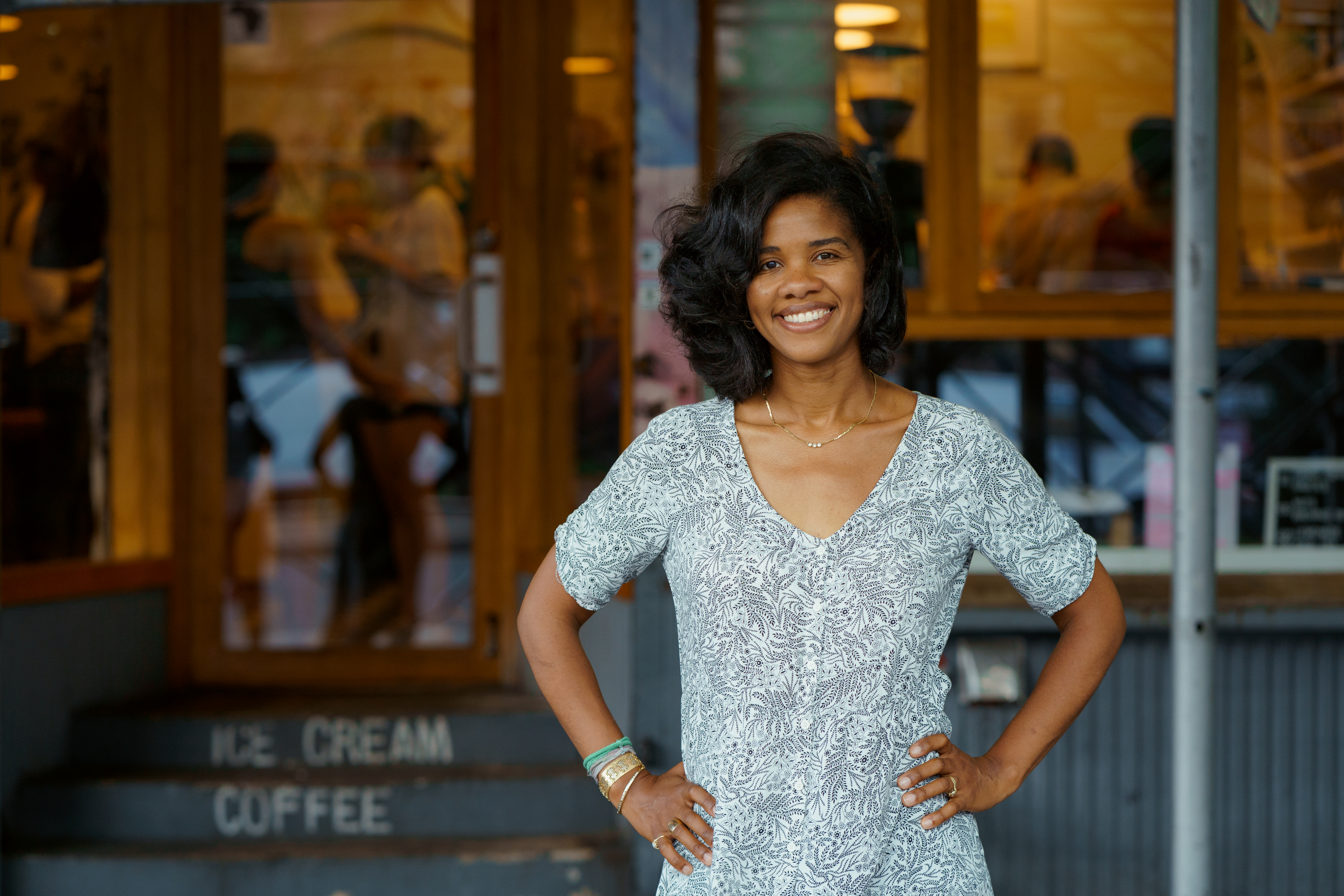 A photograph by Ajene Farrar features Petrushka Bazin Larsen, co-founder of Sugar Hill Creamery, standing confidently with her hands on her hips in front of her establishment. She is smiling and wearing a light blue and white patterned V-neck dress. The shop's entrance is visible behind her, with steps labeled "ICE CREAM" and "COFFEE," capturing her role as a leading entrepreneur and "culture maker" in the Harlem community.