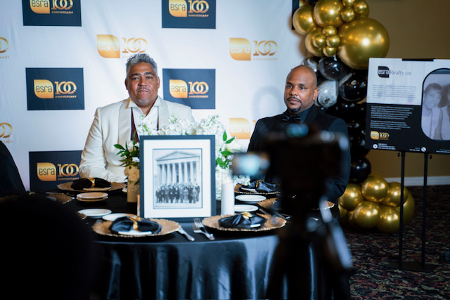 Maurice Russell Grey and Aden Seraile, 3rd generation owners of ESRA Realty, seated at their 100th Anniversary Gala in Harlem. A framed historical photograph of their predecessors is featured on the table, symbolizing the firm's century-long legacy in Black-owned real estate.