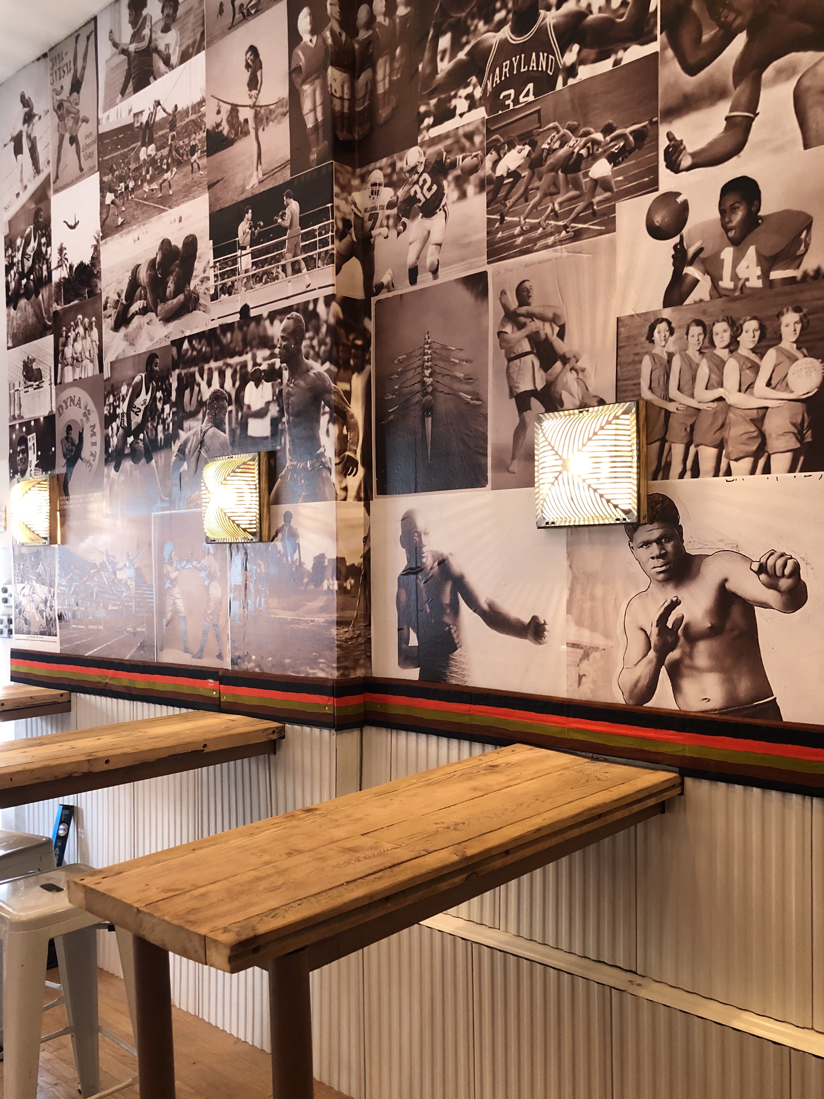 Interior of Ouaga Sports Bar in Harlem, NY, featuring a large wall mural of historic Black athletes and sports figures, wooden high-top tables, and modern gold sconce lighting.