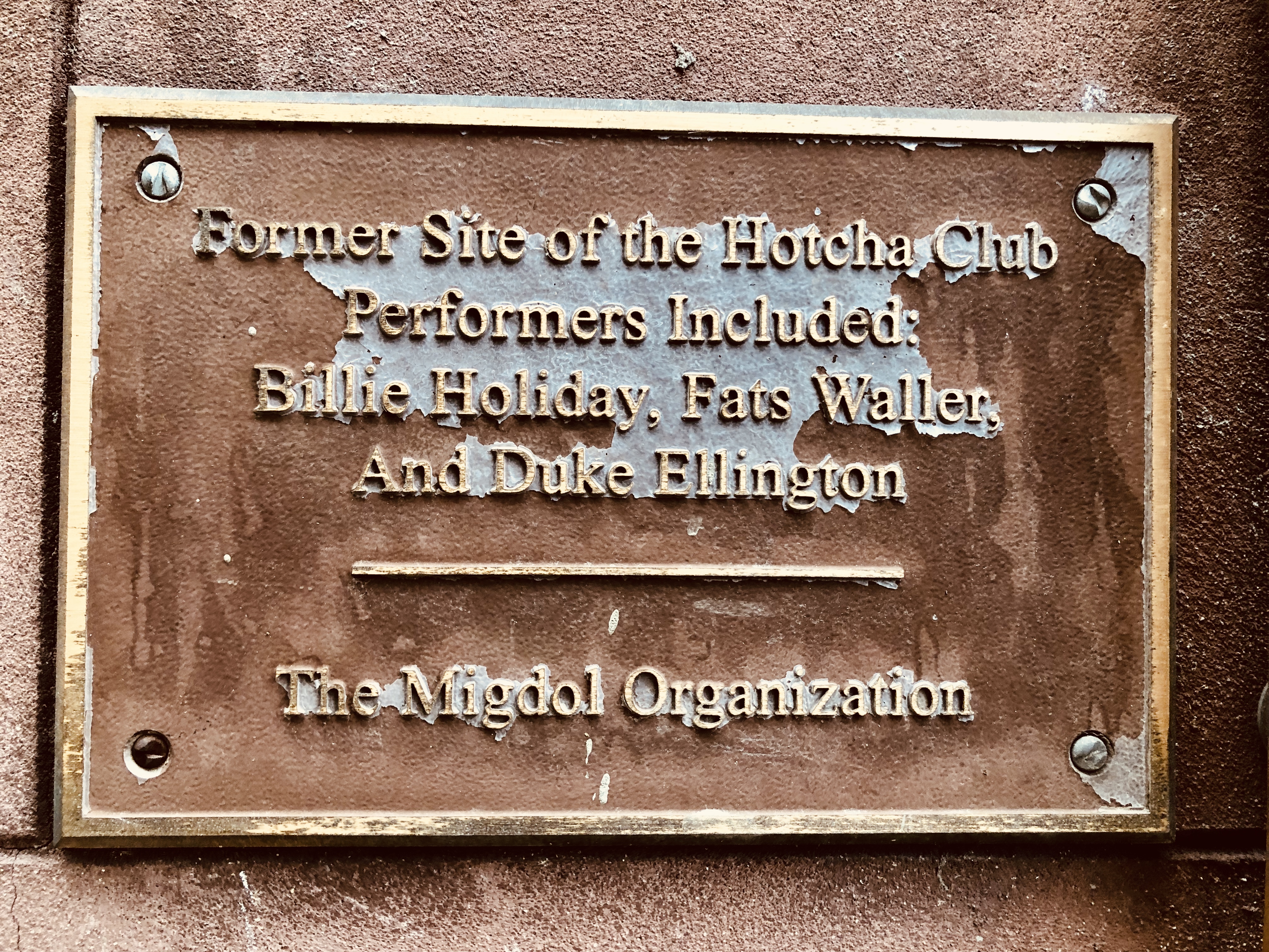 Bronze historical plaque on the exterior of Ouaga Sports Bar in Harlem, NY, identifying the building as the former site of the Hotcha Club where legends Billie Holiday, Fats Waller, and Duke Ellington performed.
