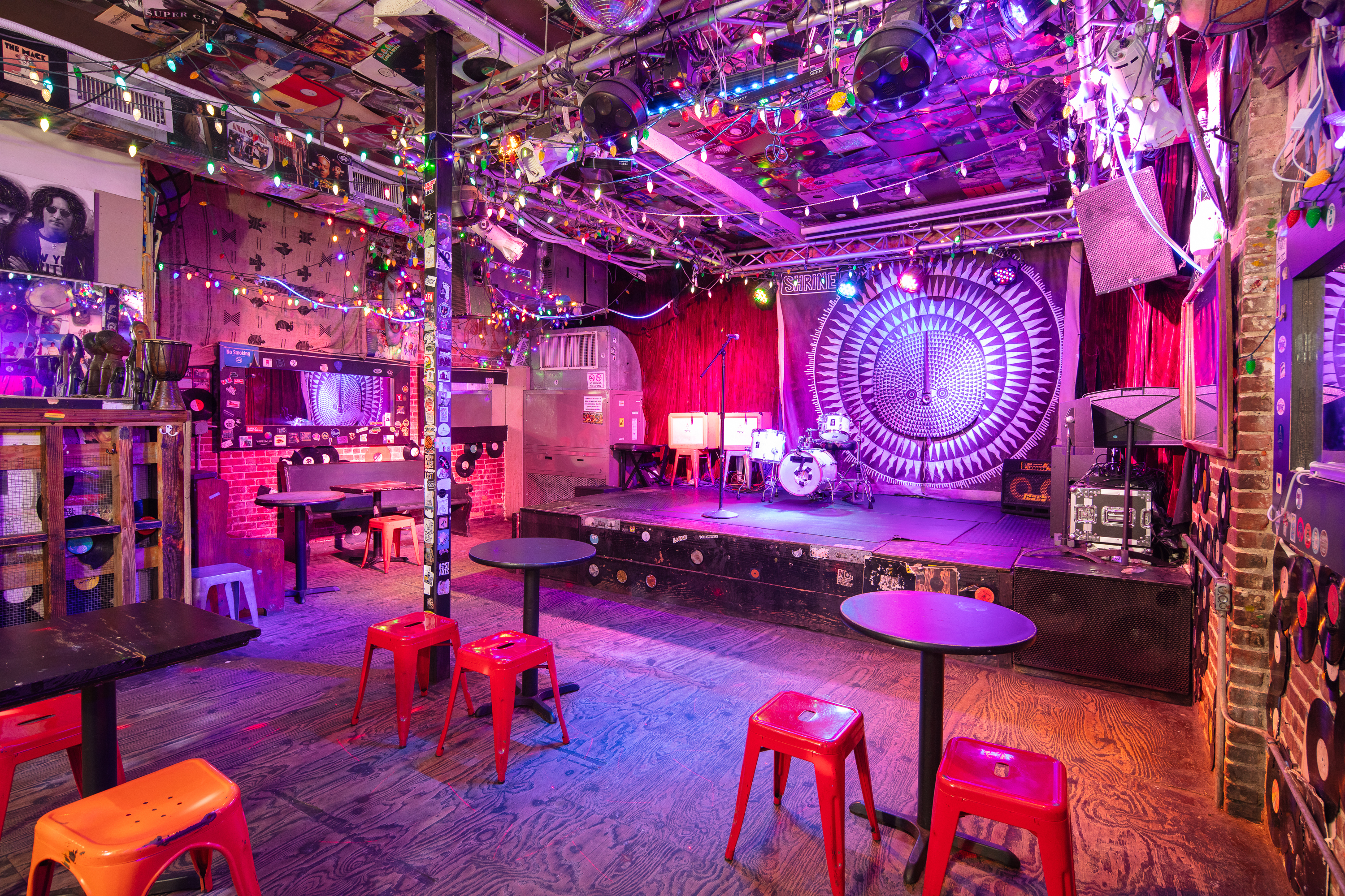 The interior of Shrine World Music Venue in Harlem, NY, featuring an empty performance stage with a drum kit, vibrant purple lighting, eclectic ceiling decor made of vinyl records, and intimate café seating with red and orange stools.