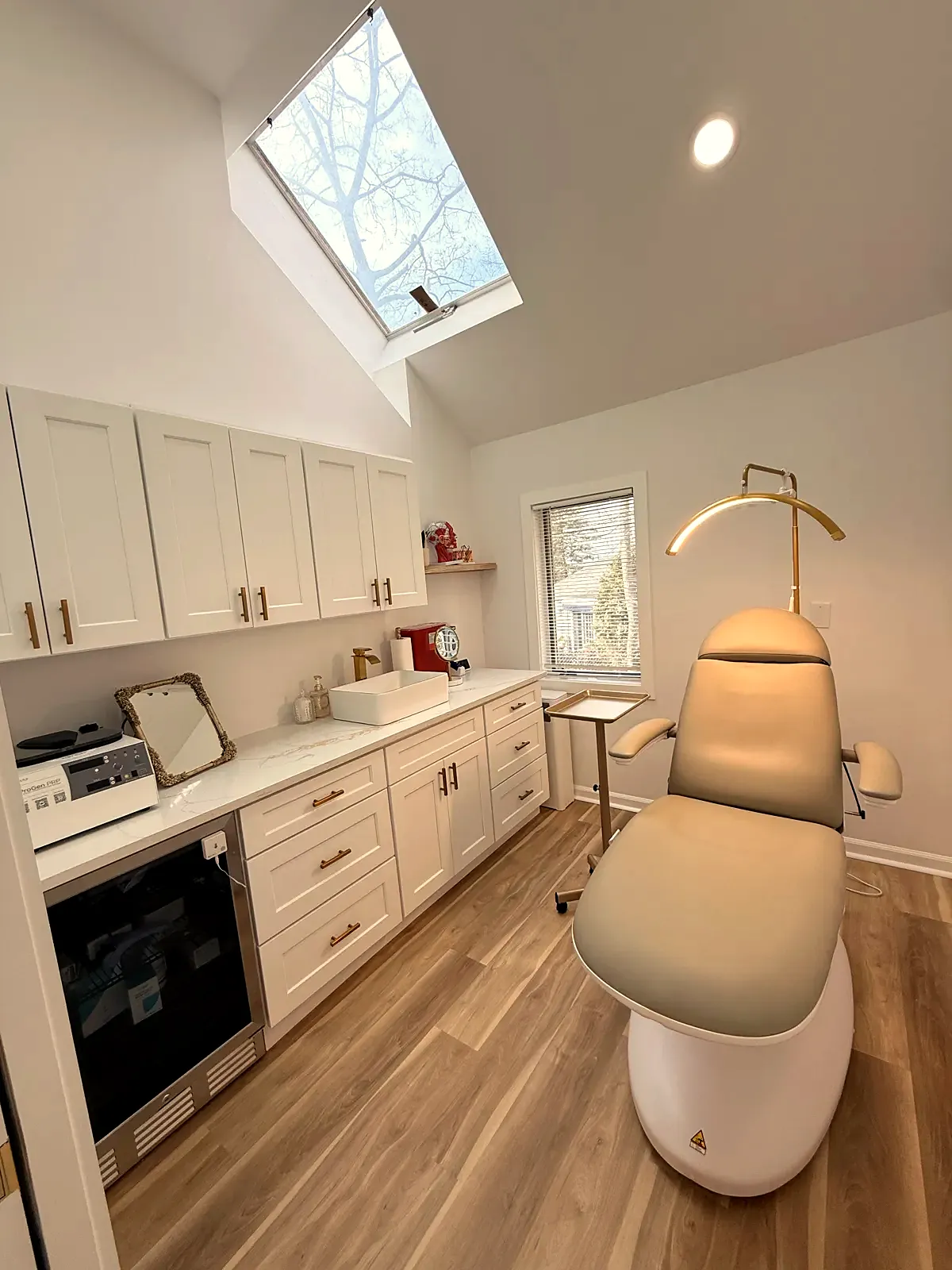 Treatment room at JENNVIE Med Spa in Westport, CT with a skylight, beige treatment chair and white cabinets with gold hardware