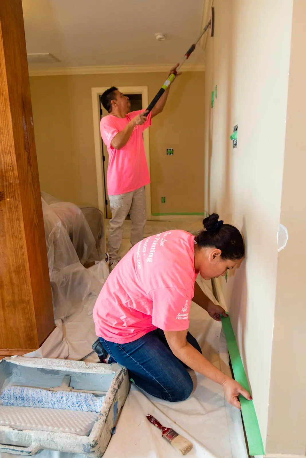 Painters taping baseboards and rolling a high interior wall