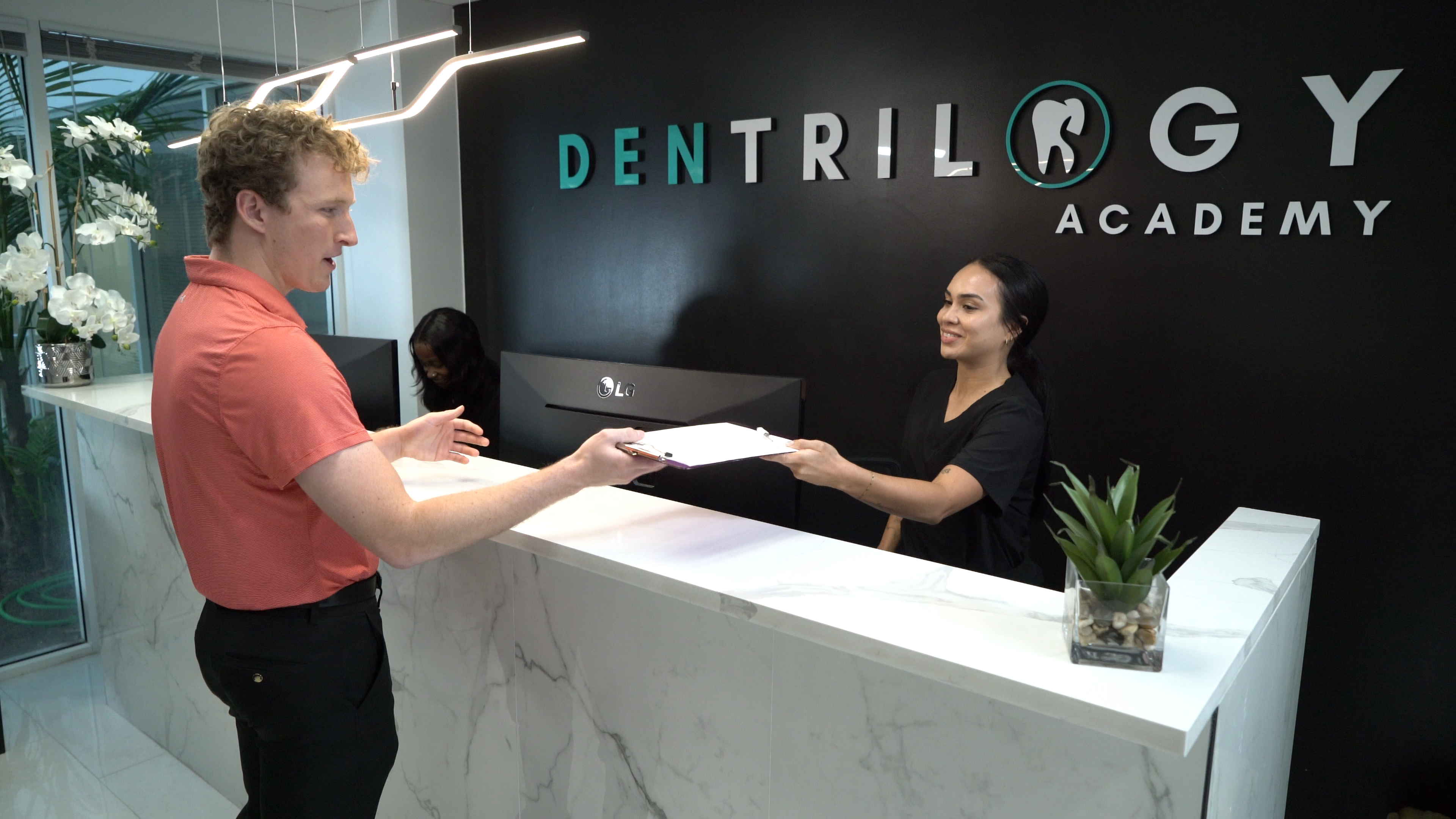 Two people practice a clipboard handoff at Dentrilogy Academy's front desk.