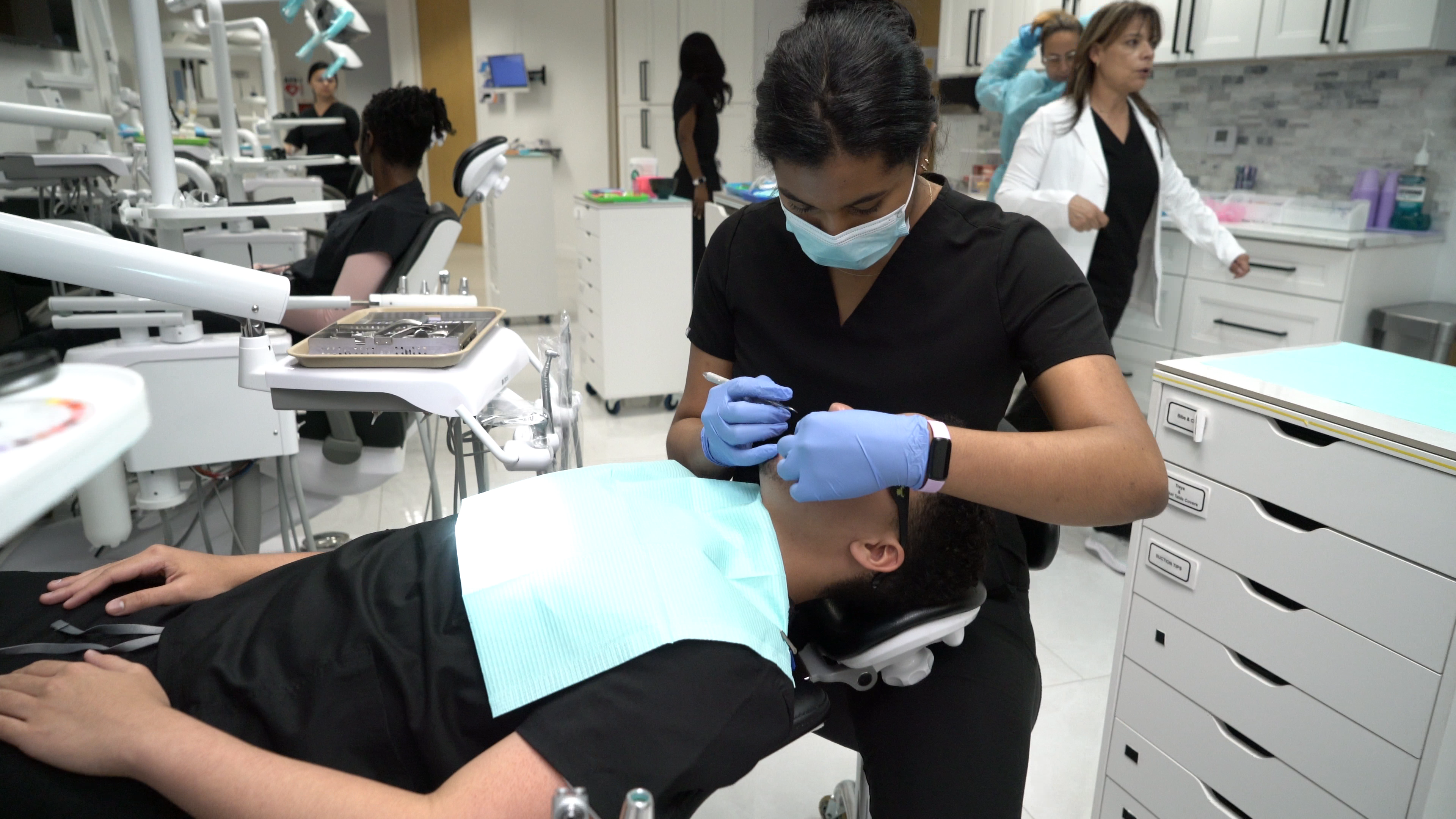 A gloved student practices chairside with instructor guidance in Dentrilogy Academy's clinical training room.