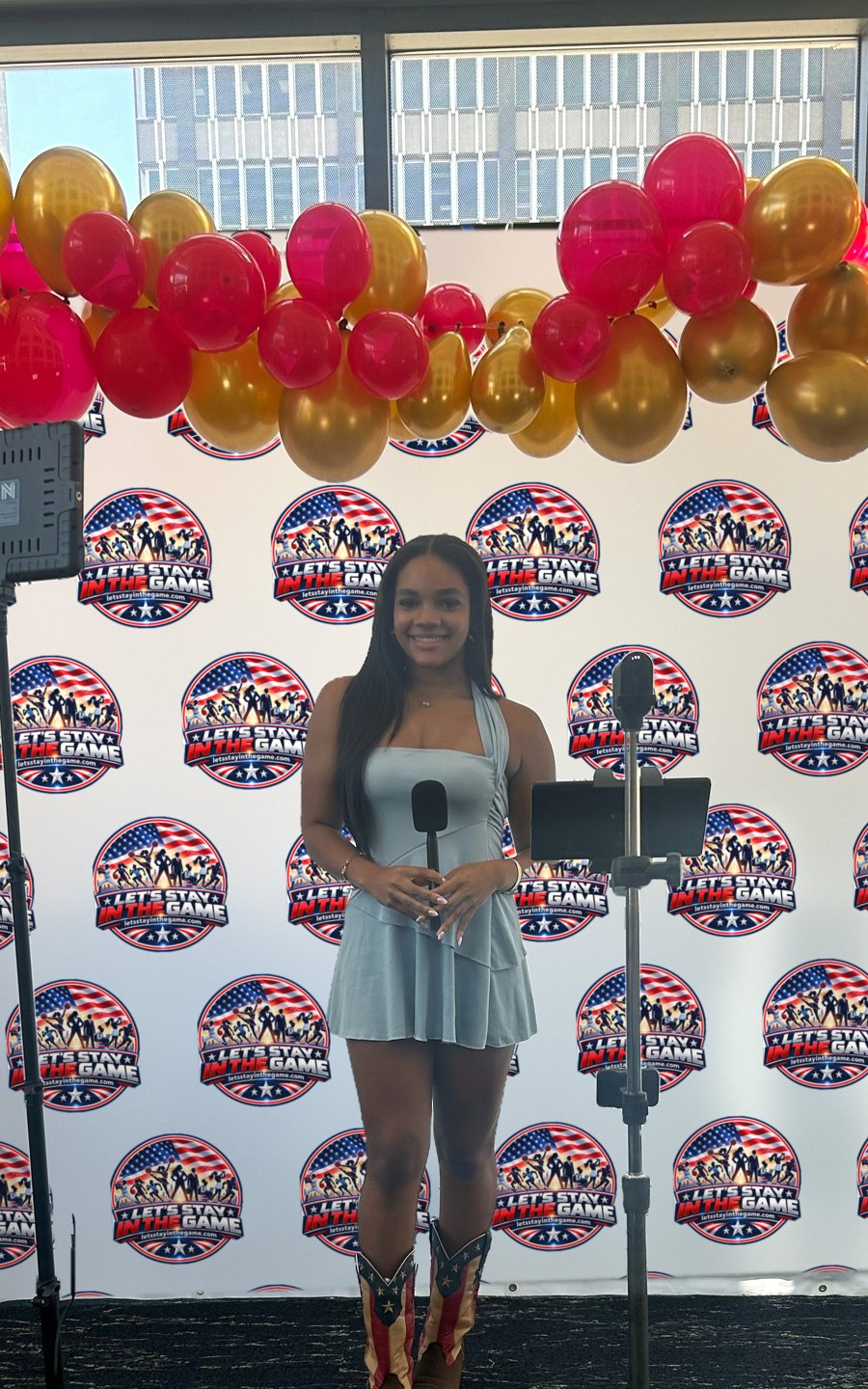 Chandler Mason holding a microphone in front of a step and repeat wall of Let's Stay in the Game badges