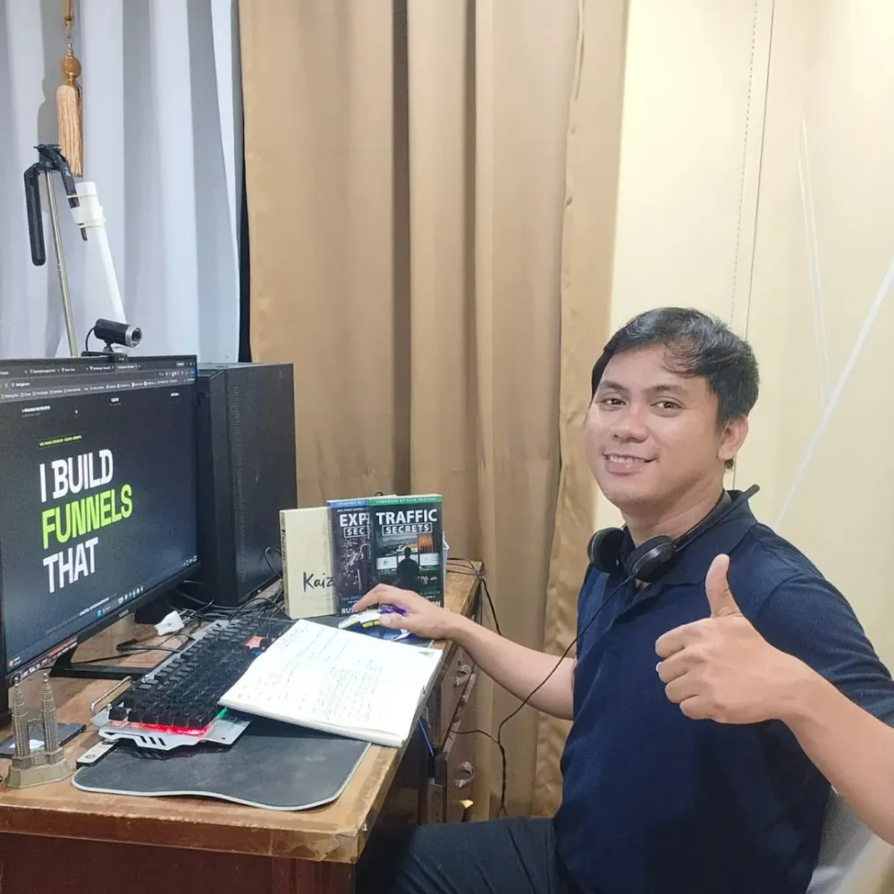 Gio at his home desk giving a thumbs up, with a headset, notebook, marketing books and a funnel page on his monitor