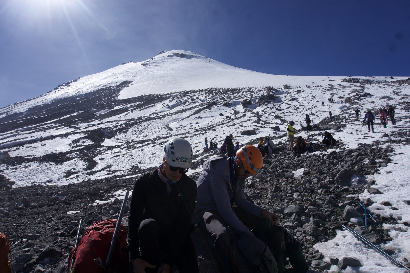 Climbers resting below the snow line on Pico de Orizaba, with the snow-covered summit dome above and bare volcanic rock below