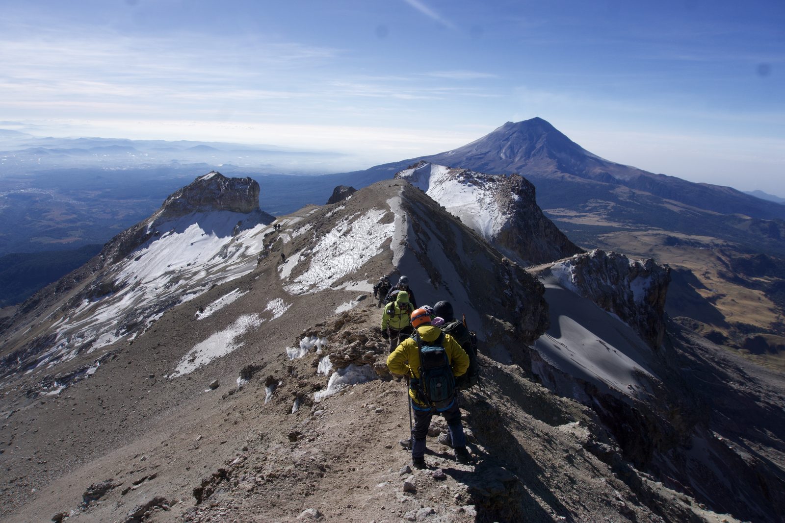 Climbers walking the Iztaccihuatl summit ridge in patchy snow with the Popocatepetl volcano rising behind them