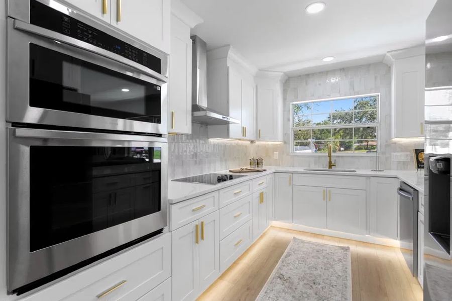 Finished Dreamway kitchen with white shaker cabinets, gold hardware, stainless wall ovens and tile backsplash