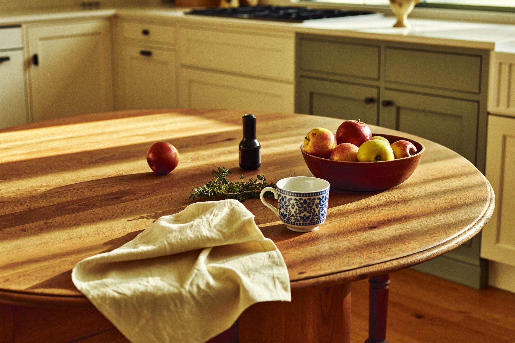 A kitchen table laid with a mug, apples and herbs in warm light