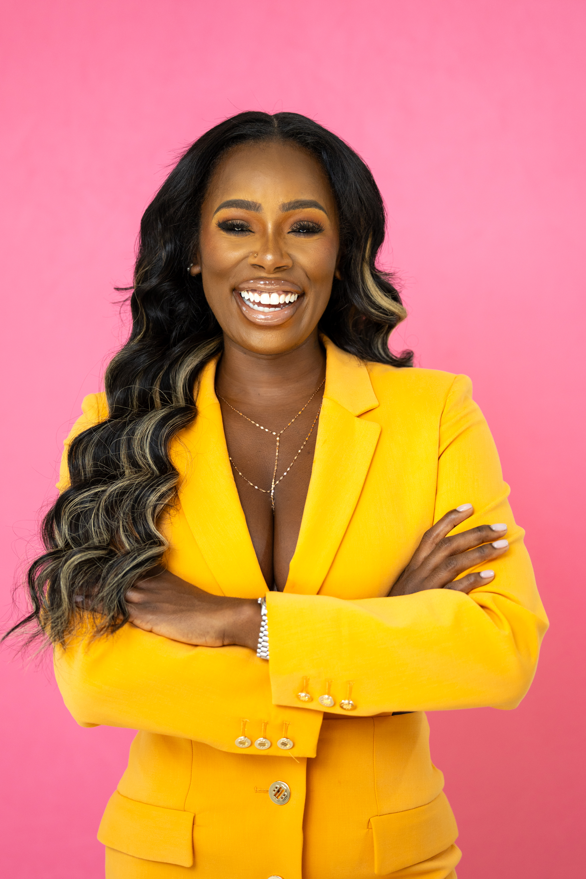 Quellyn Rosa Kennedy , black woman smiling radiantly in a yellow blazer, posed against a pink background, showcasing her vibrant energy and confidence.