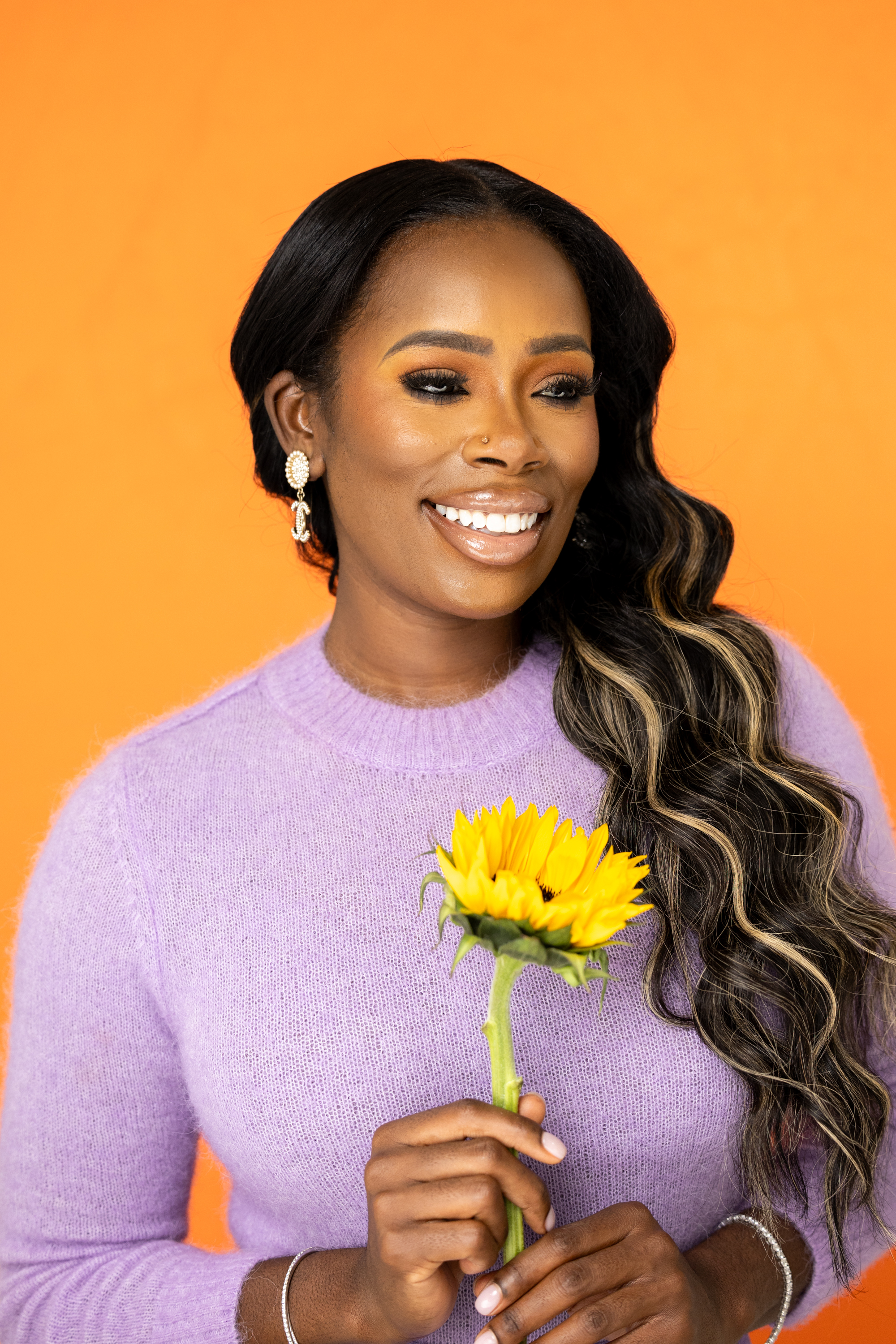 Quellyn Rosa Kennedy black woman smiling gracefully in a lavender sweater, holding a vibrant sunflower against a bold orange background, radiating warmth and positivity.