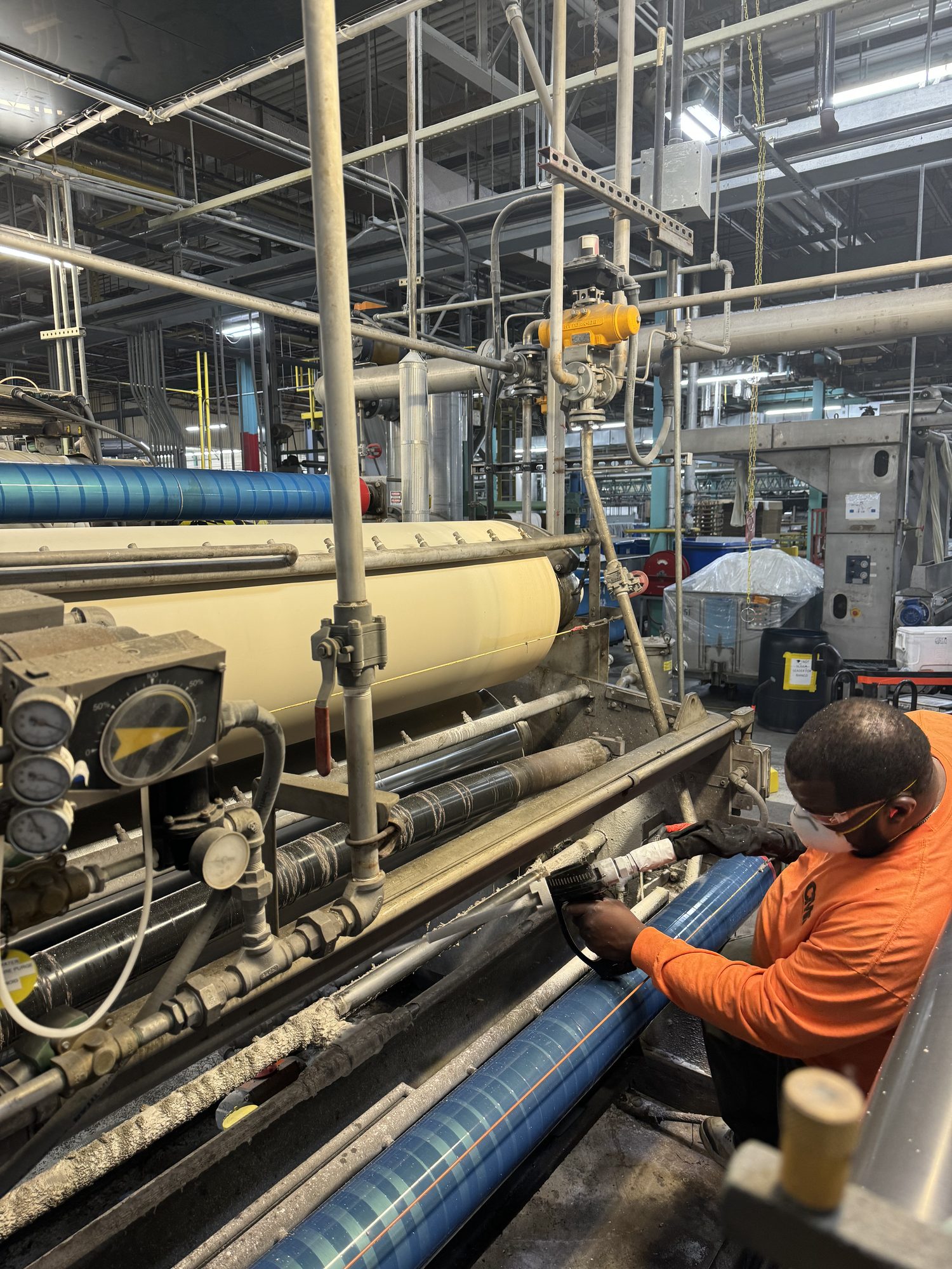 Tech in respirator and orange shirt dry ice blasts a textile roller without pulling it from the line.