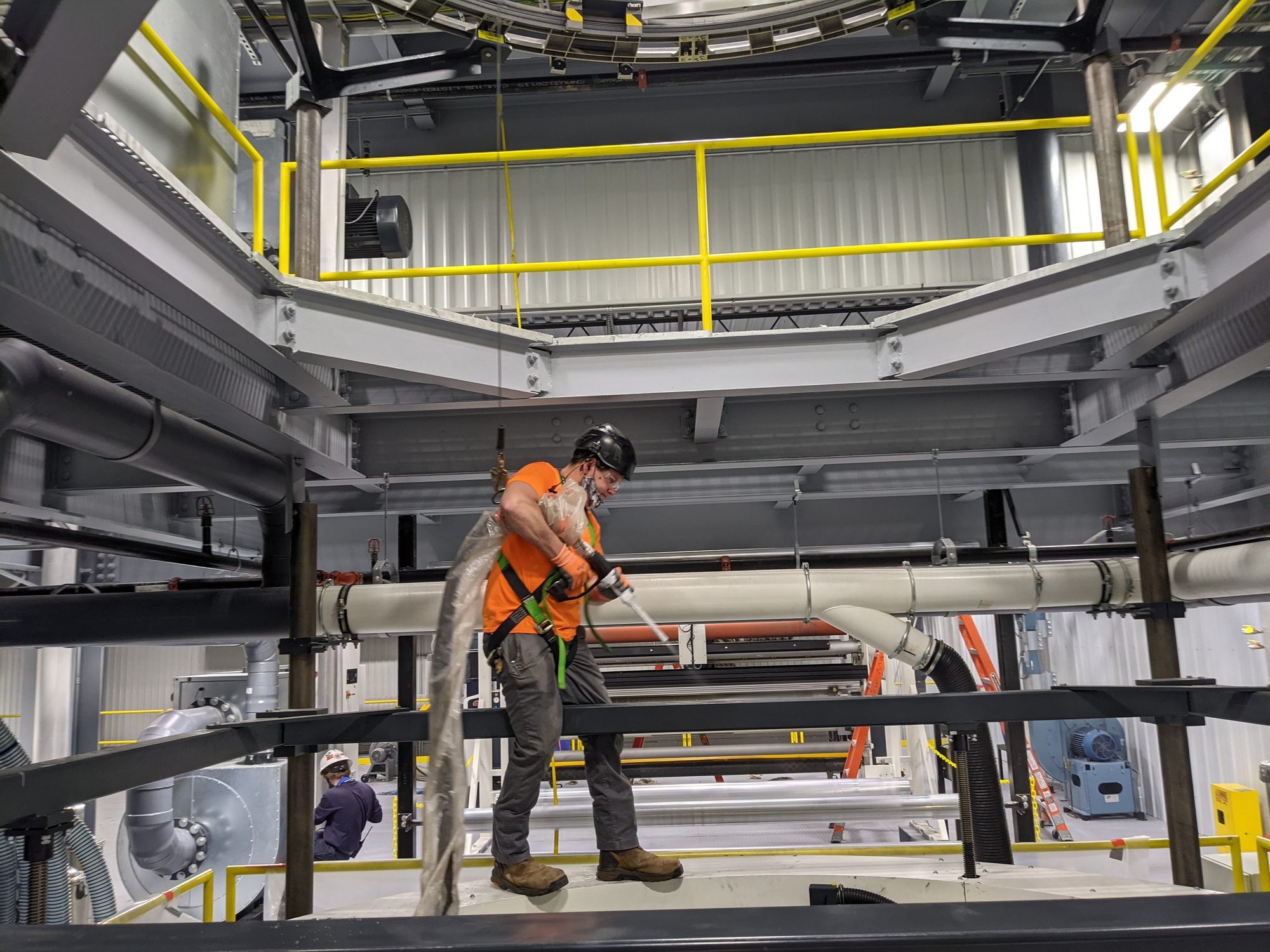Harnessed and tied-off tech dry ice blasts machine framework from the top deck of a film line.