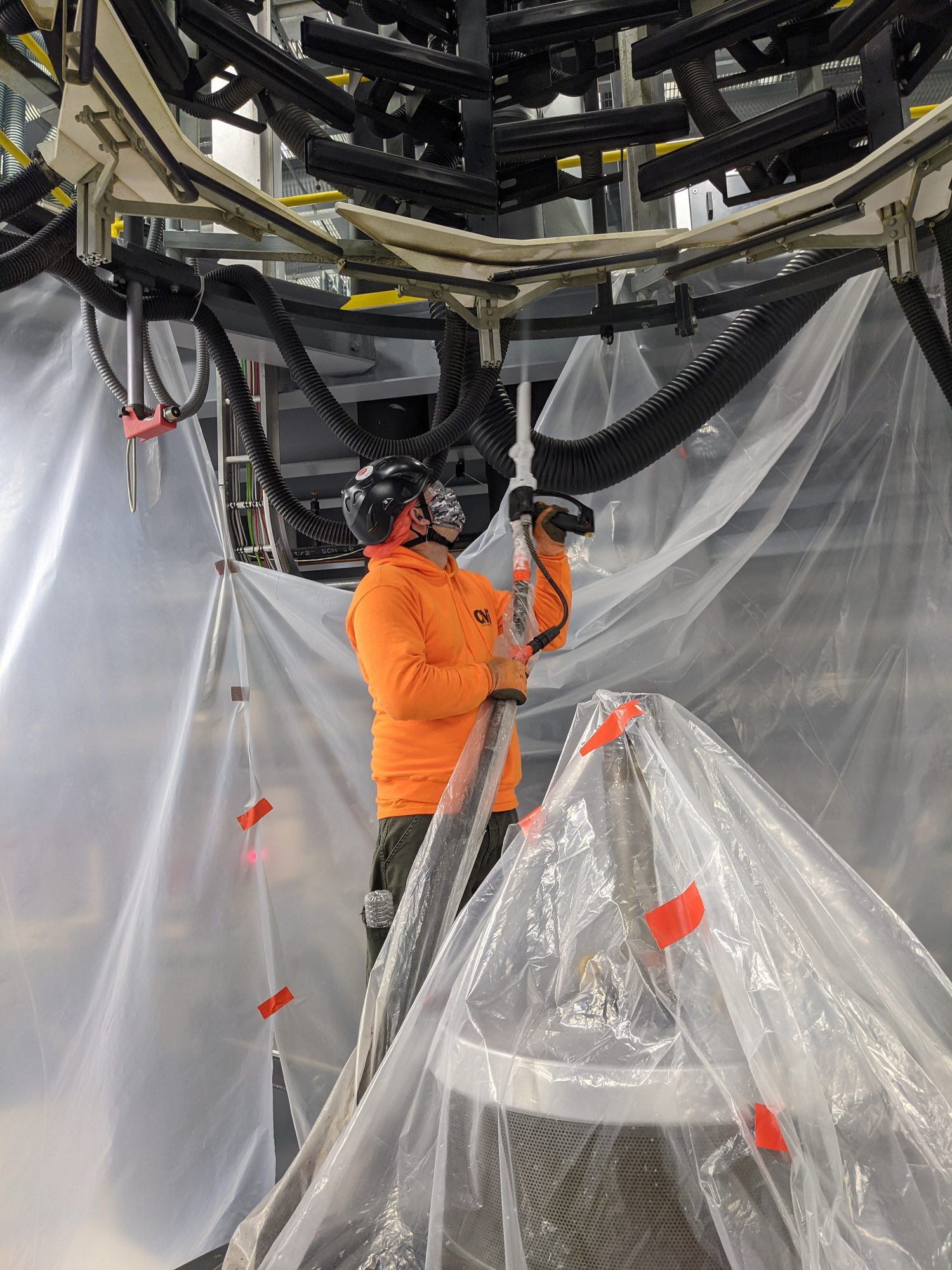 Tech in a CMI hoodie reaches overhead with the blast gun to clean a cooling ring inside containment.