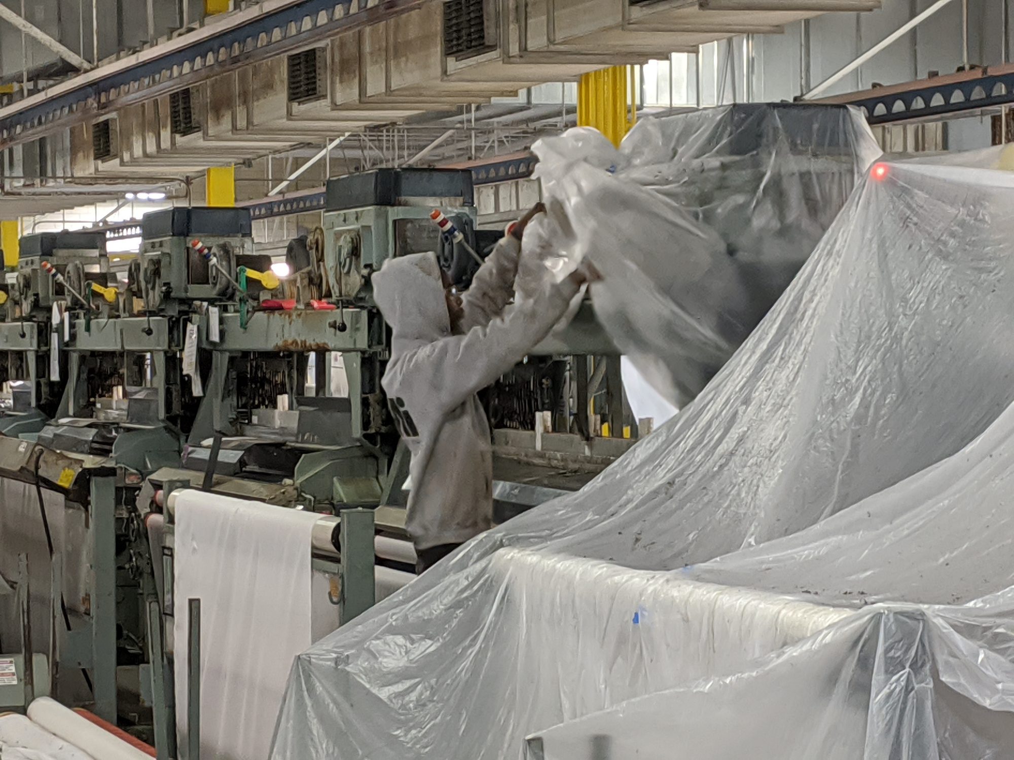 Tech wraps textile looms in plastic sheeting to protect them before shutdown cleaning.