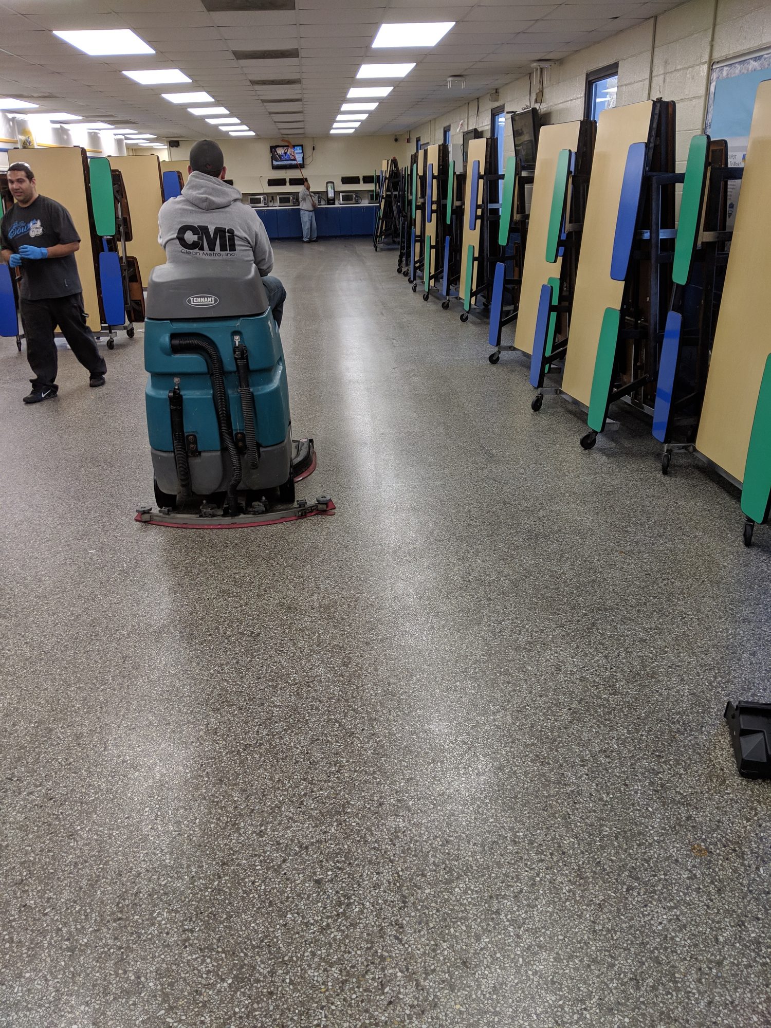 CMI tech runs a ride-on scrubber down a long cafeteria aisle.