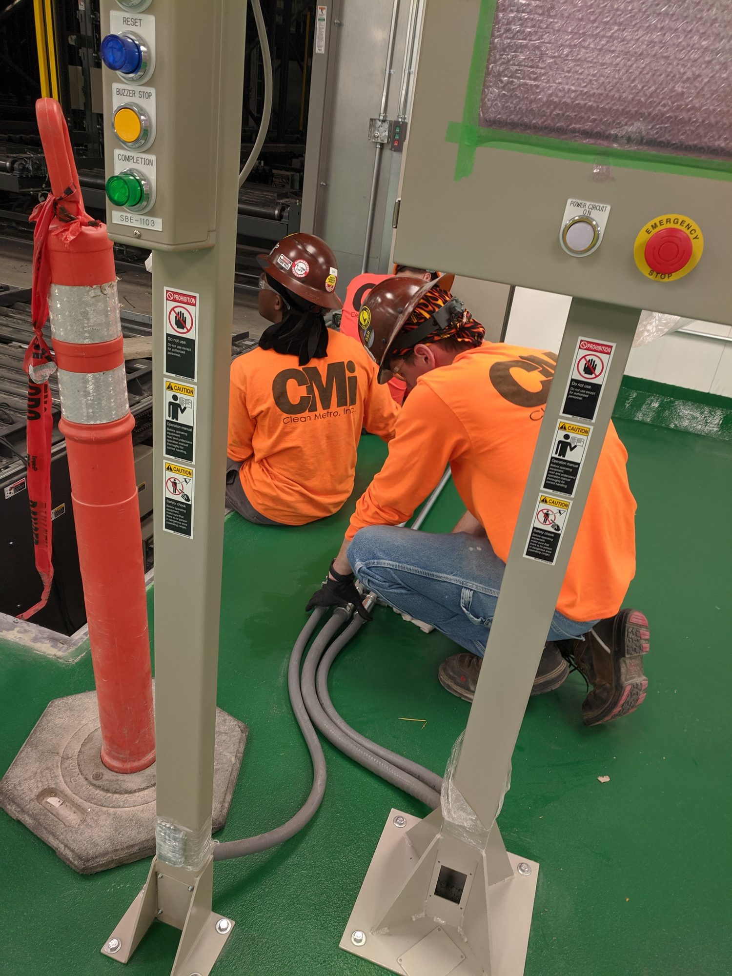 Two CMI techs kneel to vacuum debris from under new production equipment on a green epoxy floor.