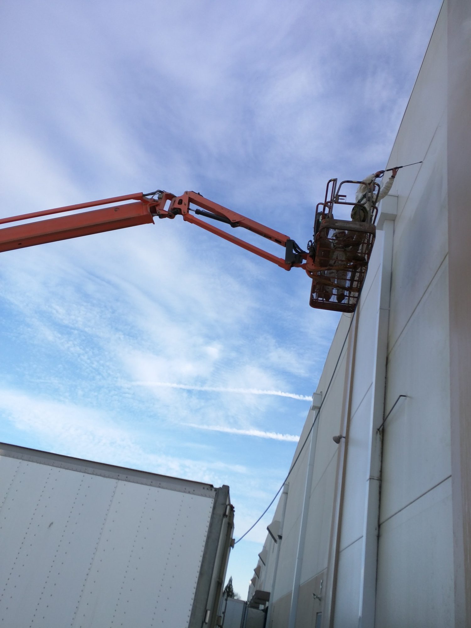 Boom lift holds a wash tech at the top of a 40-foot warehouse wall between parked trailers.