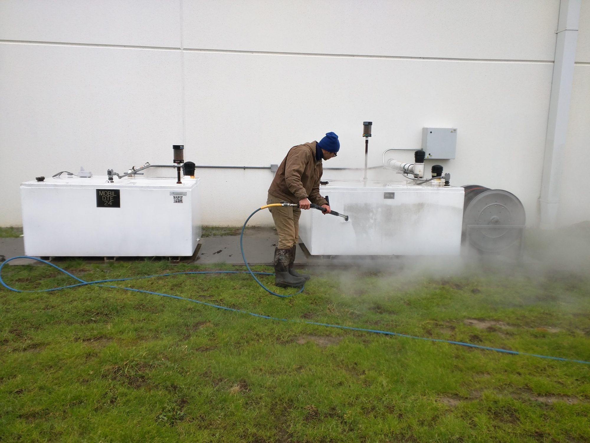 Tech hot-water washes outdoor equipment tanks against the building, steam rolling off the surface.