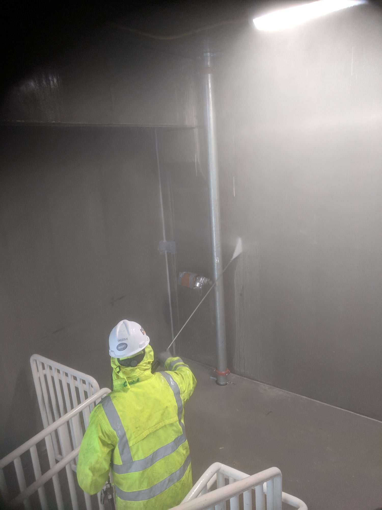 Tech in rain gear pressure washes a new parking-deck stairwell, spray mist filling the shaft.