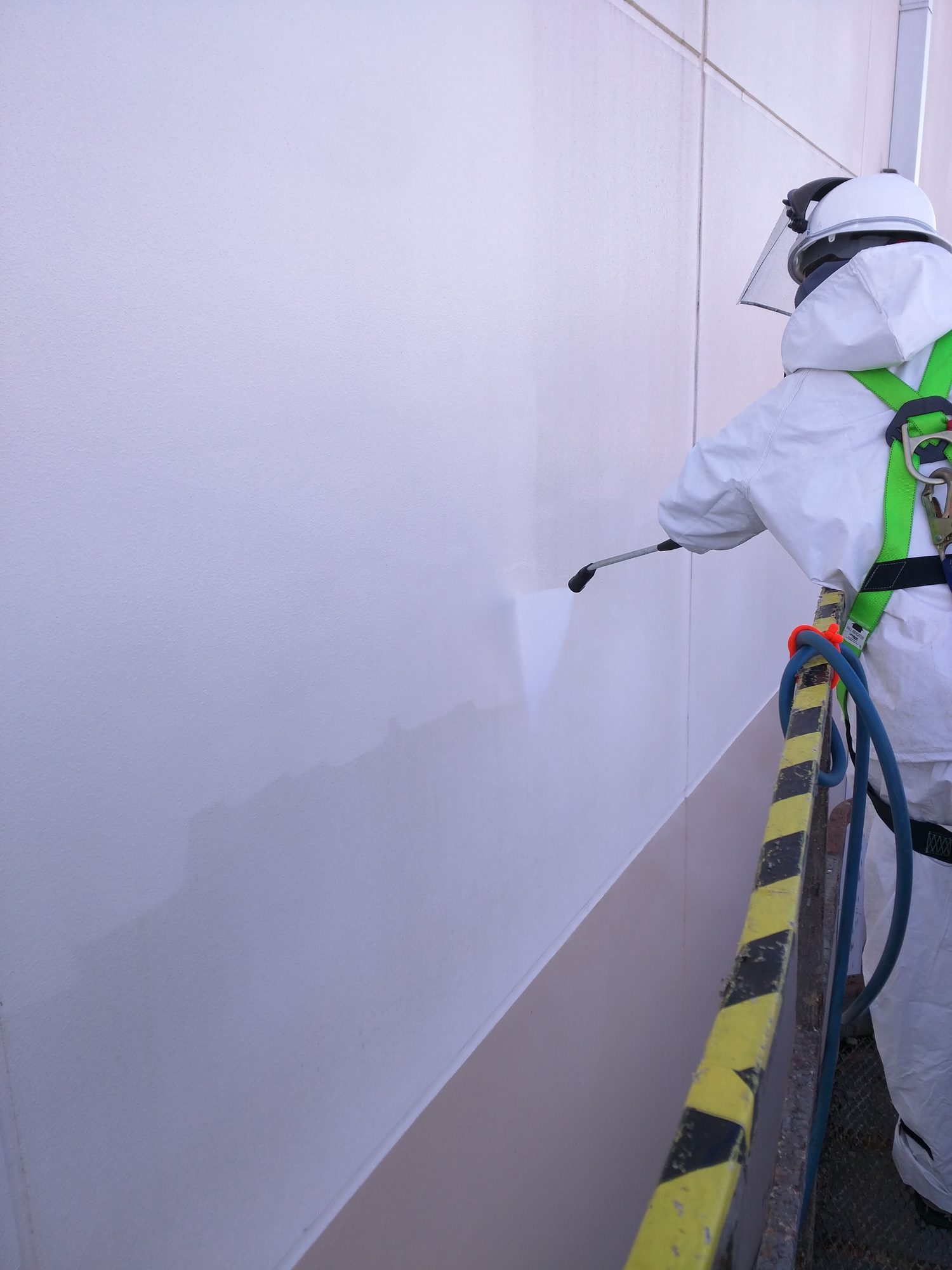 Tech in Tyvek and harness runs a wash wand down a white warehouse wall, leaving a bright clean band.